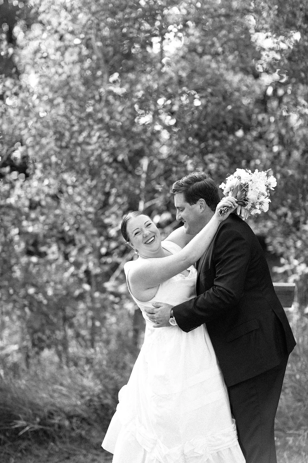 A bride and groom dancing outdoors, smiling and embracing each other, with trees in the background in black and white.