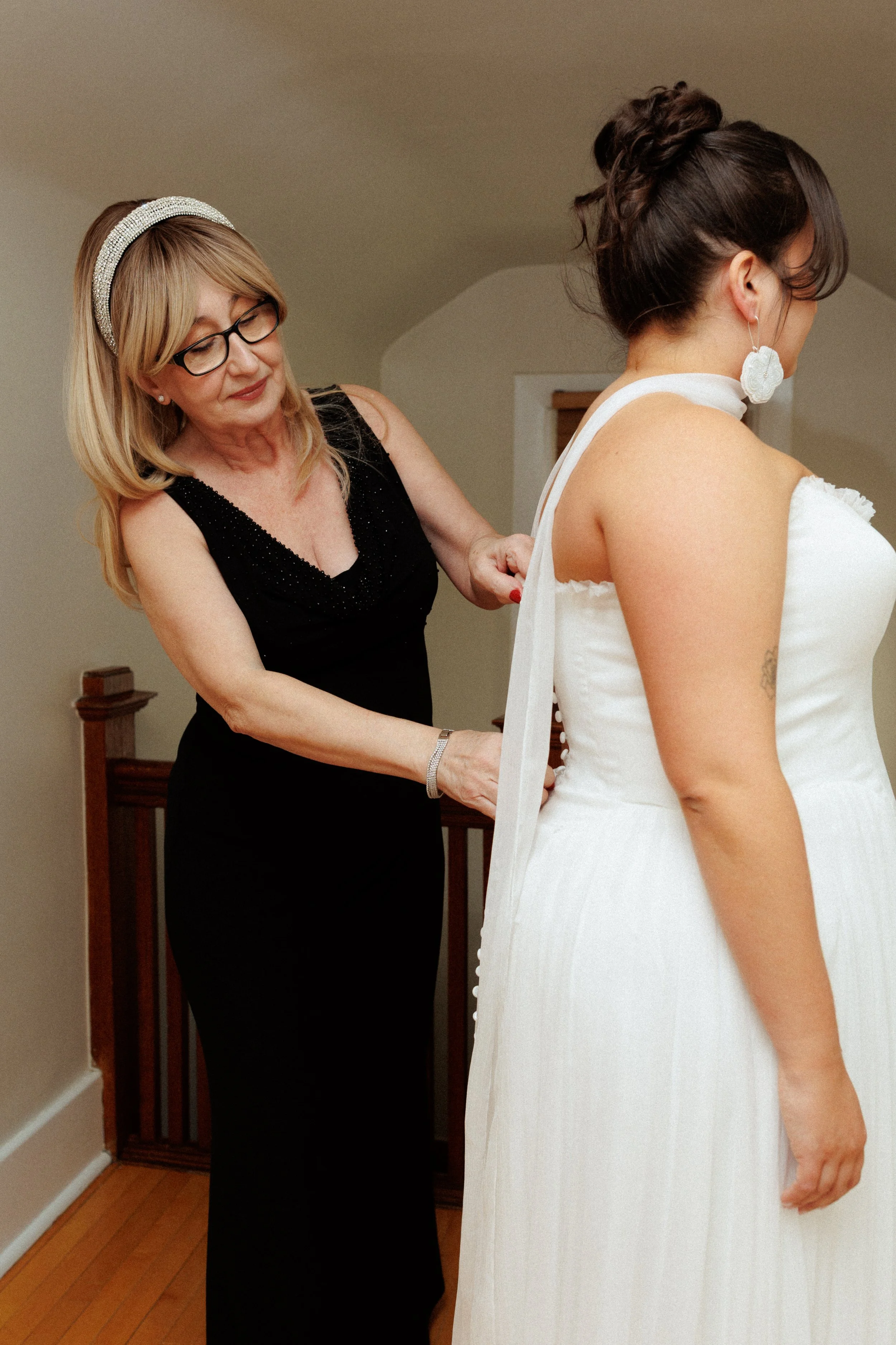 A woman helps a bride put on her wedding dress in a room with wooden flooring.