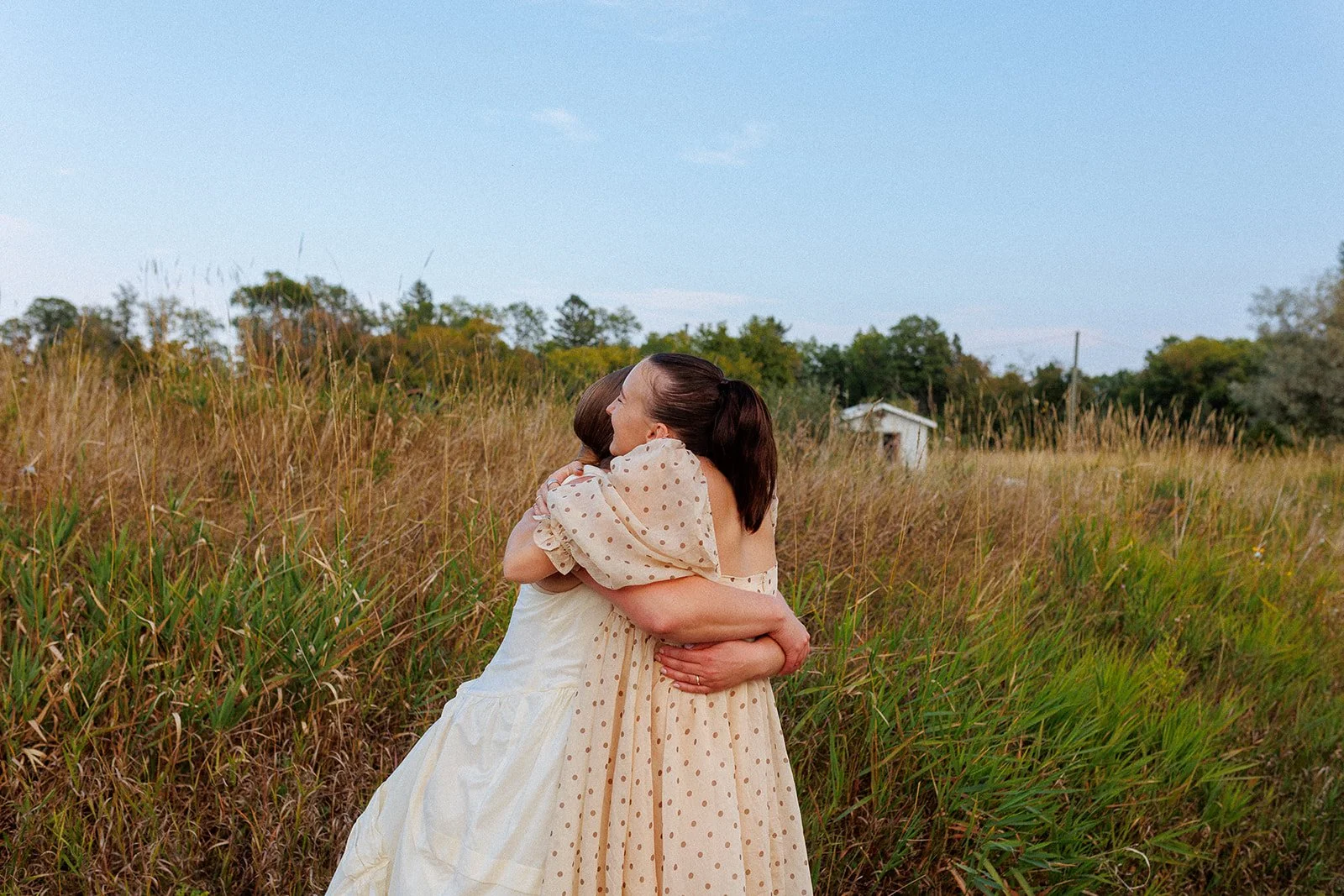 Two women hugging in a grassy field with trees and a small white structure in the background.
