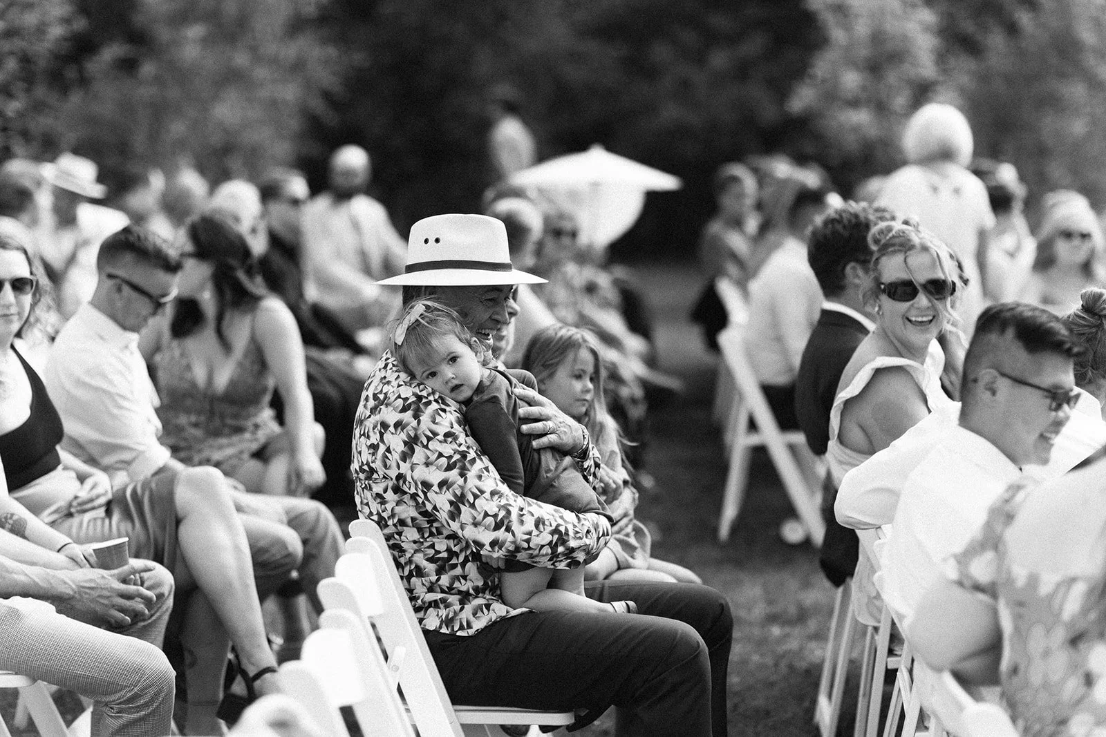 People sitting outdoors in chairs, attending an event. A woman wearing a hat is holding a child, who looks towards the camera. Multiple other people, some wearing sunglasses and casual clothing, are visible in the background, some smiling and others 