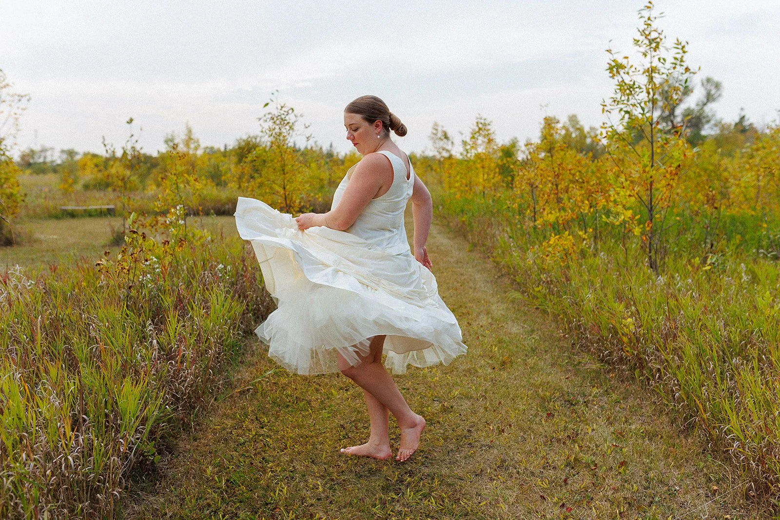Woman in a wedding dress standing barefoot on a grassy path in an outdoor setting with yellow and green trees around.
