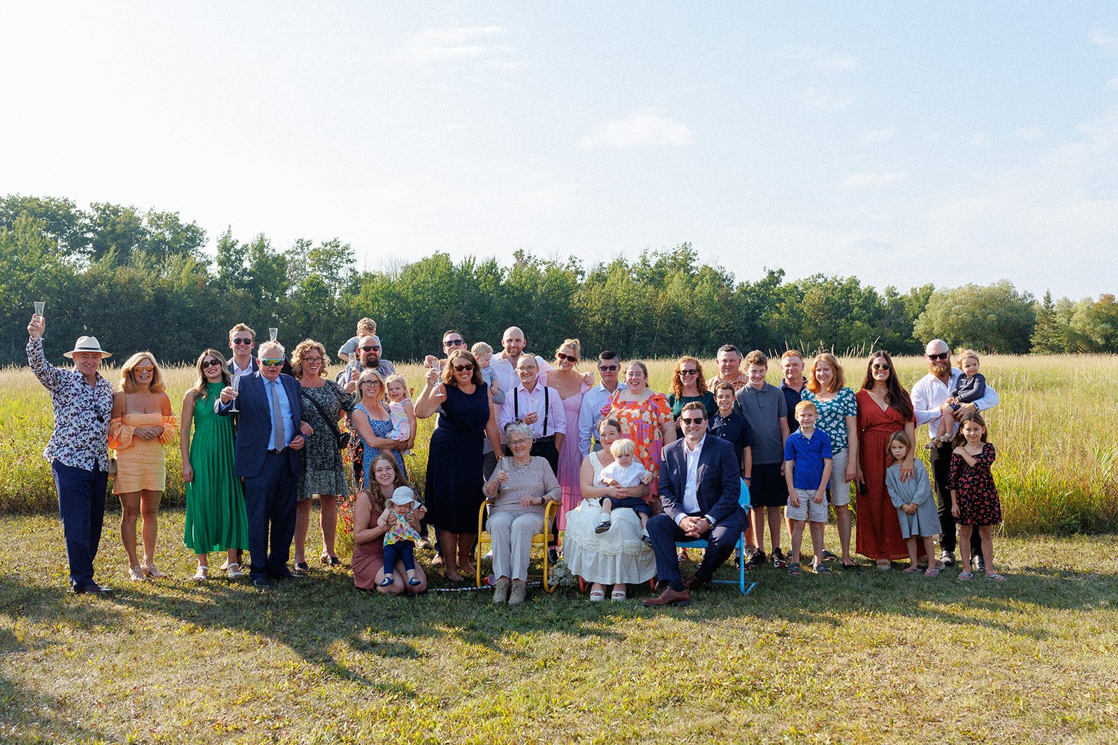 Group of people celebrating outdoors in a field on a sunny day, including children, adults, and elderly, some holding glasses of champagne.