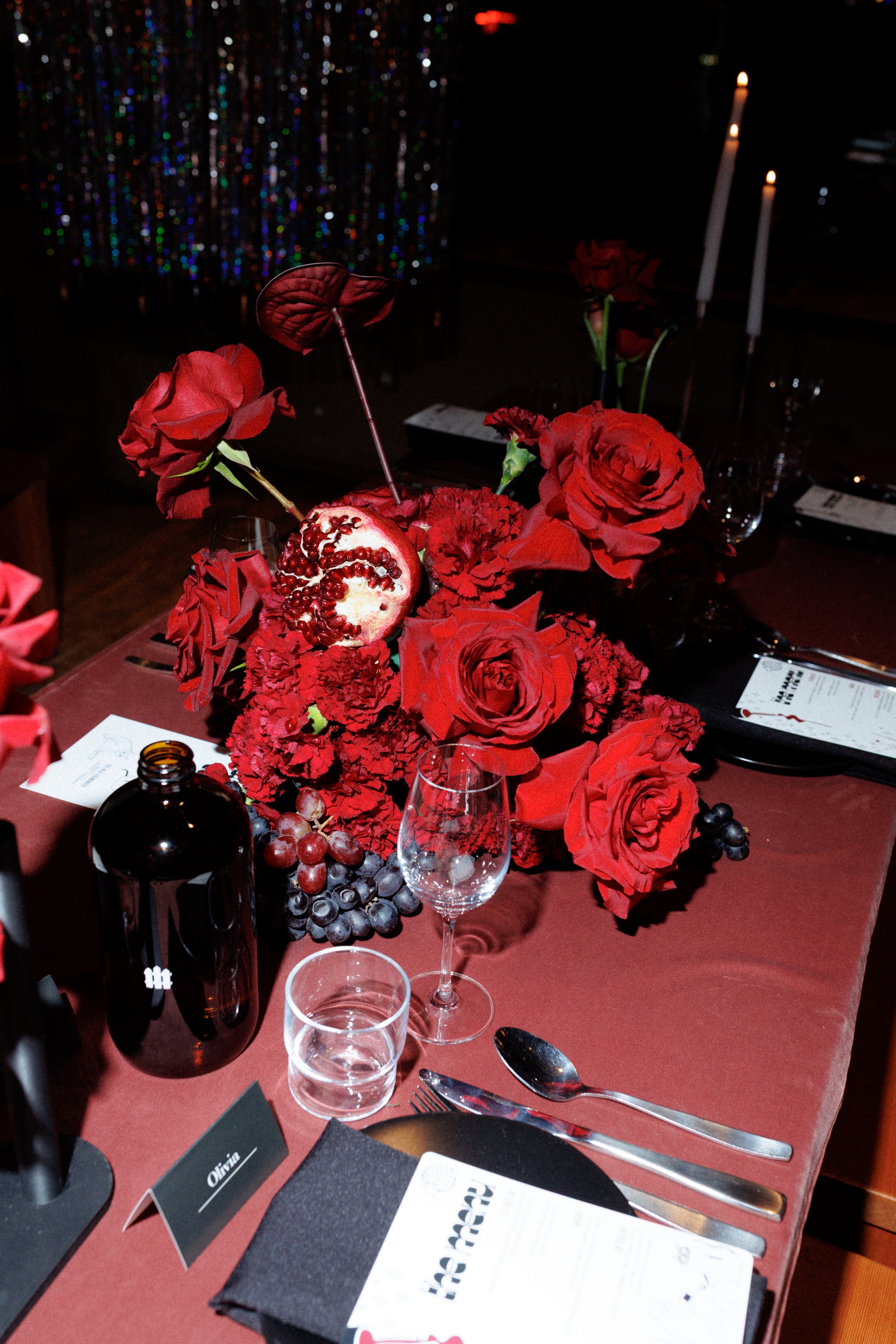 A table decorated with a large red floral centerpiece, including roses and other flowers, accented with grapes and a partially opened pomegranate, set with glassware, utensils, and a black menu card, with candles in the background.