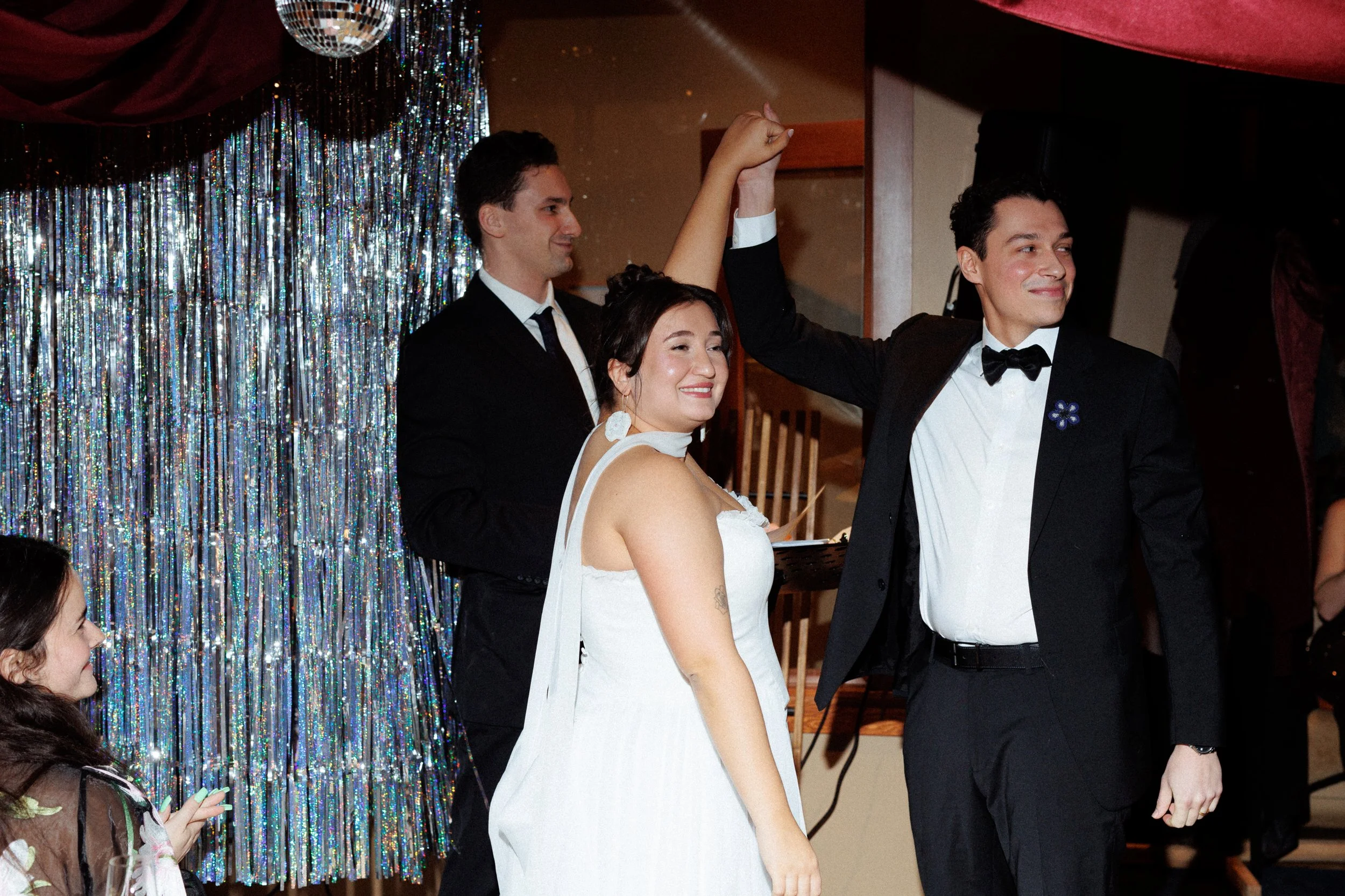 A couple dances at a wedding reception while another man in a tuxedo joins in the dance, and a young dancer in the foreground watches and smiles. The background features a shiny, silver curtain and a disco ball.