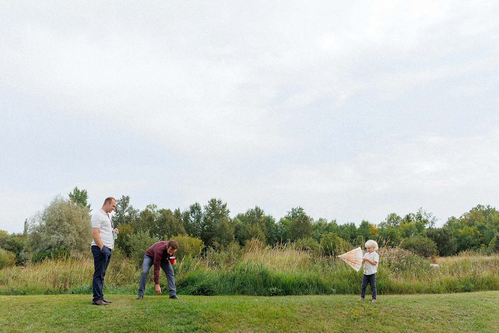 Three people in a grassy field with trees in the background, two men and a child with a paper umbrella.