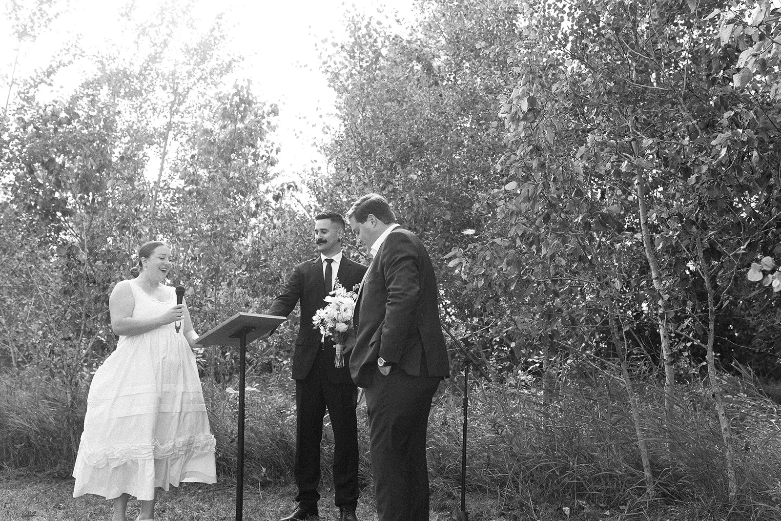 A woman in a white dress is speaking into a microphone during a wedding ceremony outdoors, with two men in suits standing beside her, one holding a bouquet of flowers, all surrounded by trees.