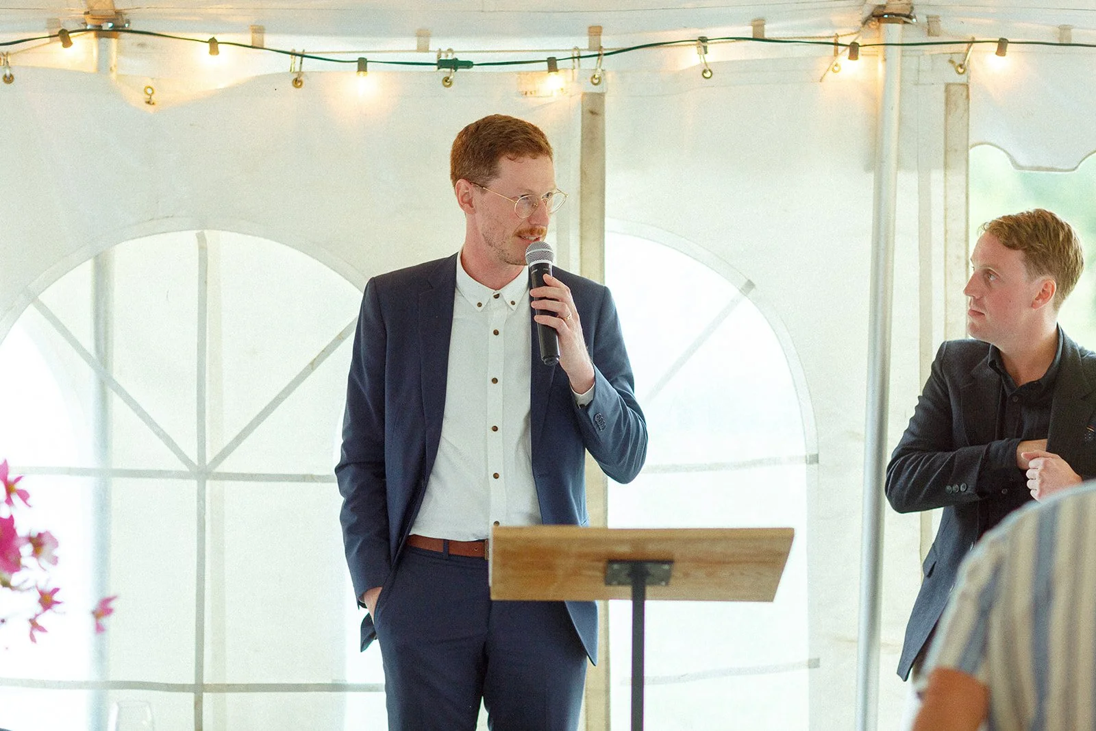 A man in a dark blue suit and glasses is speaking into a microphone at a formal event or gathering inside a tent with string lights overhead.
