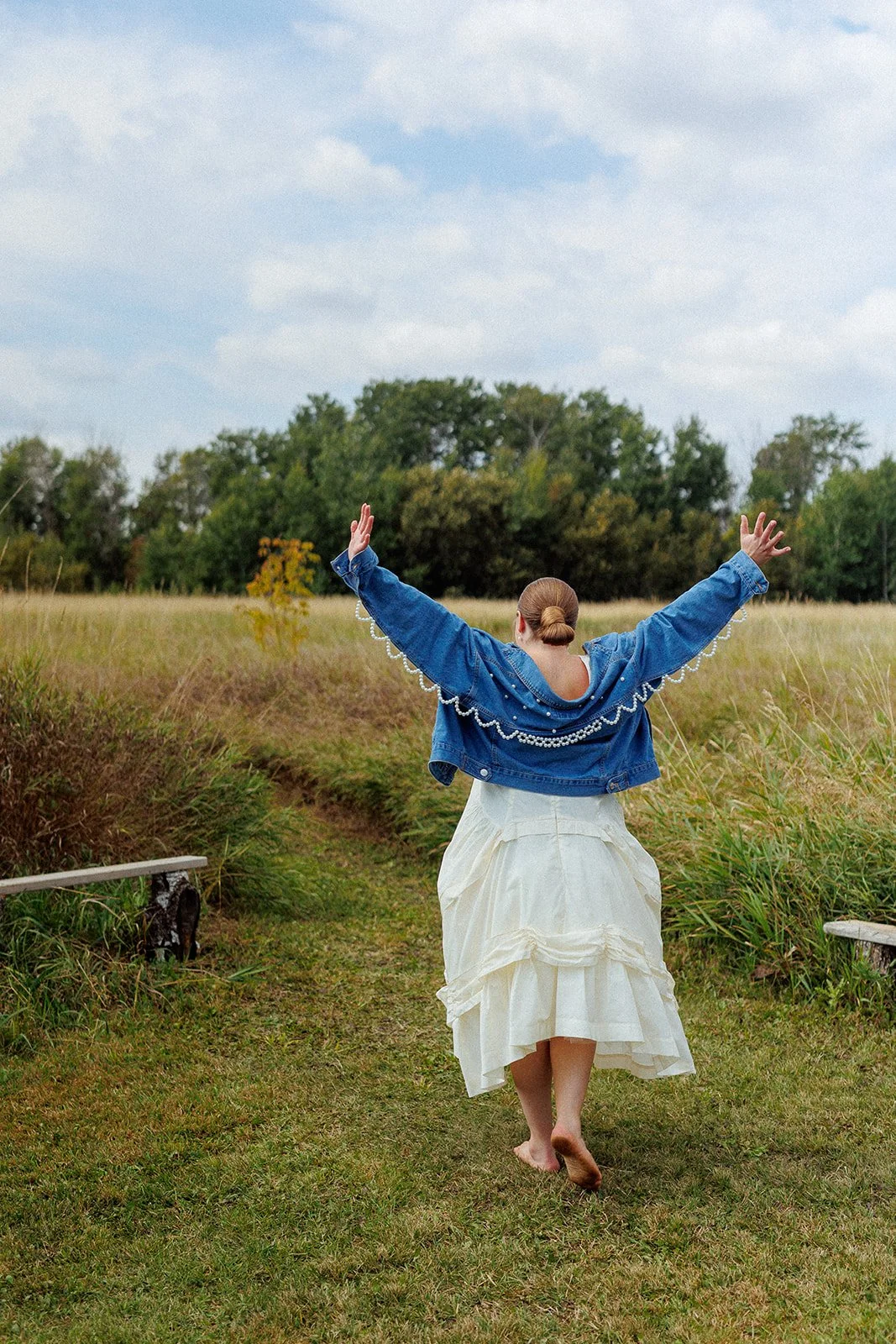 Woman in a white dress and denim jacket walking barefoot on a grassy path with arms raised, in an open field with trees in the background under a cloudy sky.