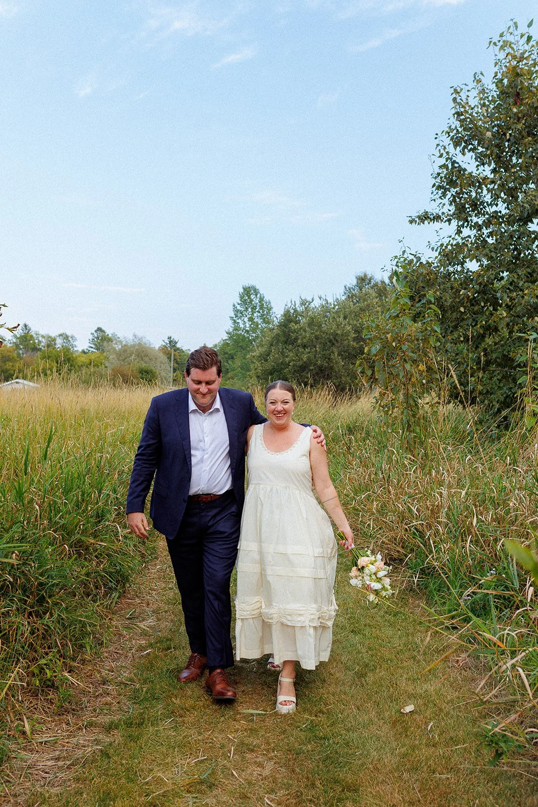 A happy couple walking arm in arm through a grassy path in a field, smiling, with the woman holding a bouquet of flowers.