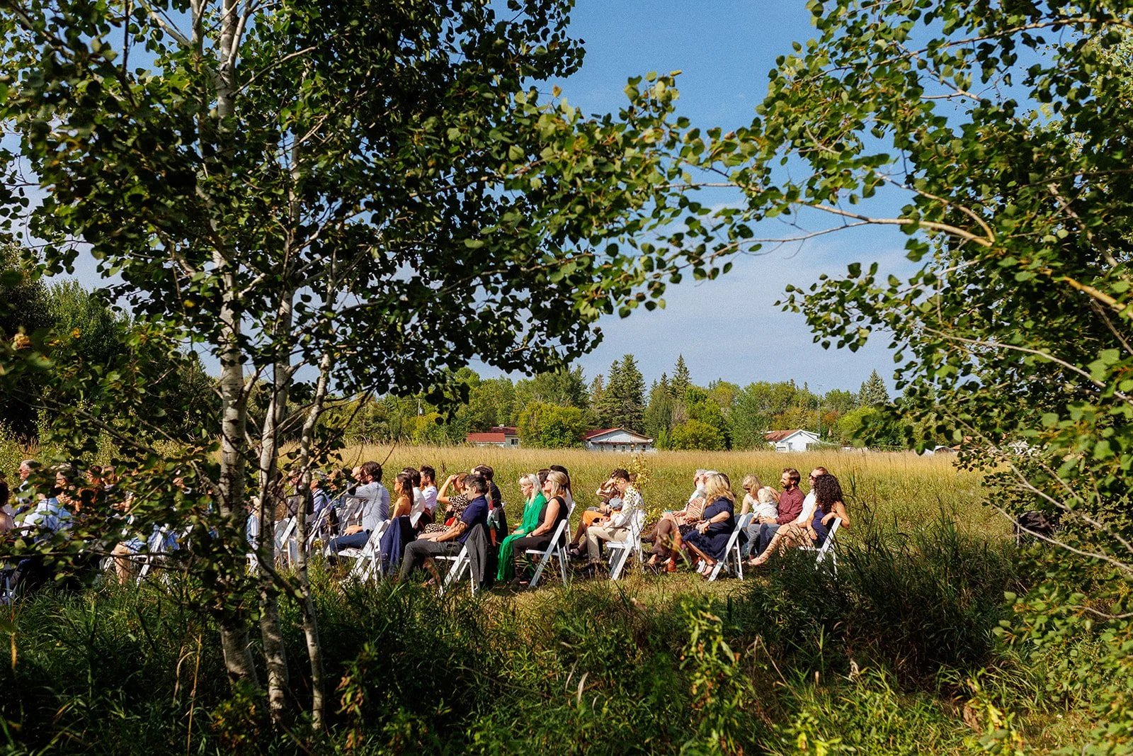 People seated outdoors on white chairs in a grassy field with trees and houses in the background, during a sunny day.