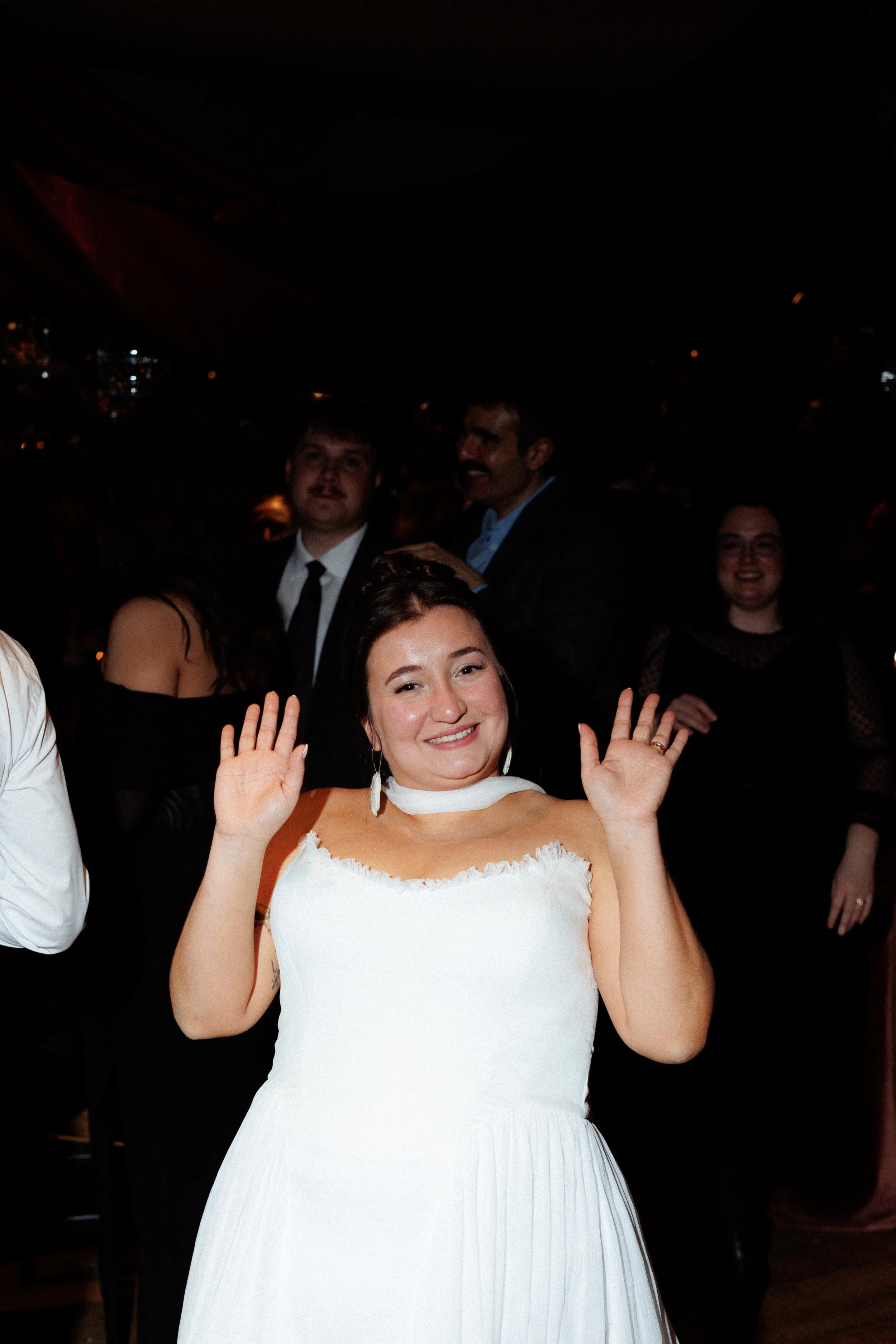 A woman in a white dress smiling and waving at a party or celebration, with other people dancing and socializing in the background.