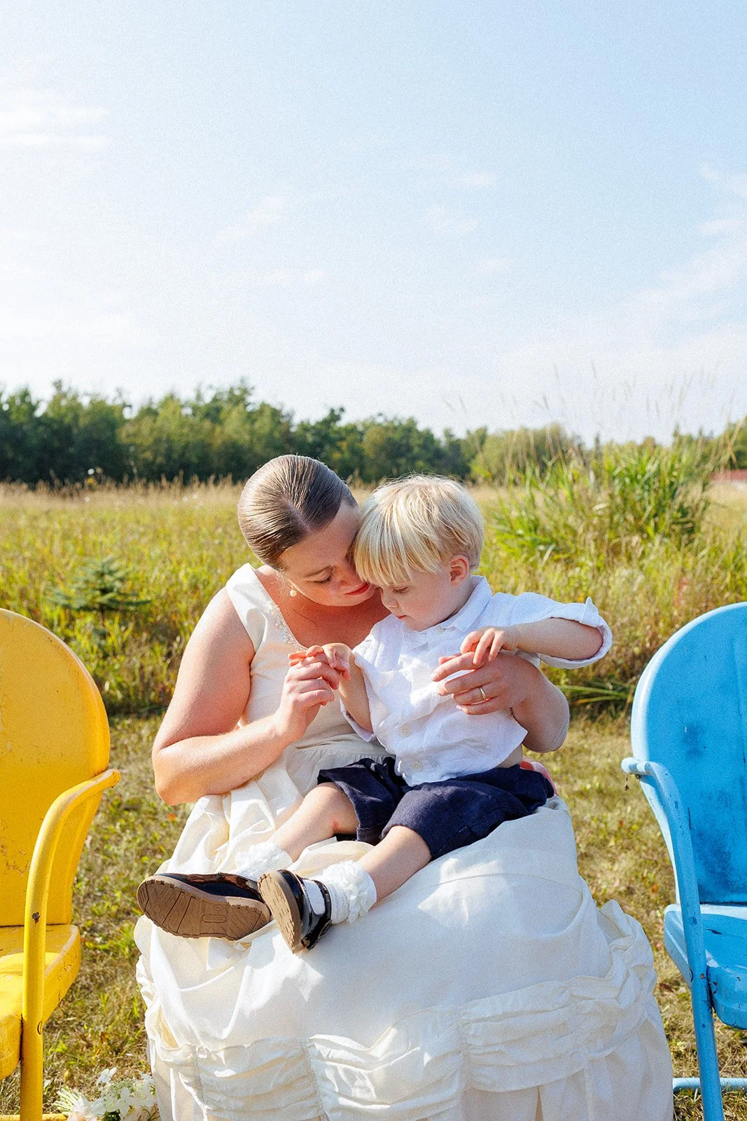 A woman and a young boy sitting on a white cloth outdoors, holding hands and looking at each other, with colorful chairs around them, in a grassy field under a blue sky.