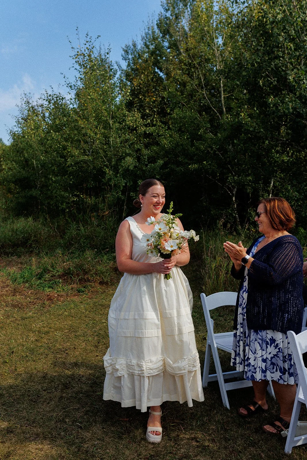 A bride holding a bouquet of flowers smiling as she is being applauded by a woman in a navy sweater and sunglasses at an outdoor wedding ceremony with green trees and grass in the background.