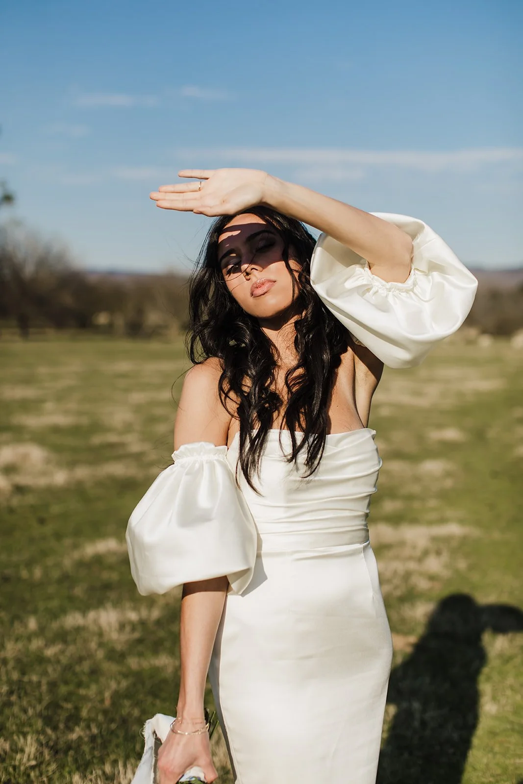 Model wearing satin gown stands in the sun in the middle of a sheep field holding a petite bouquet