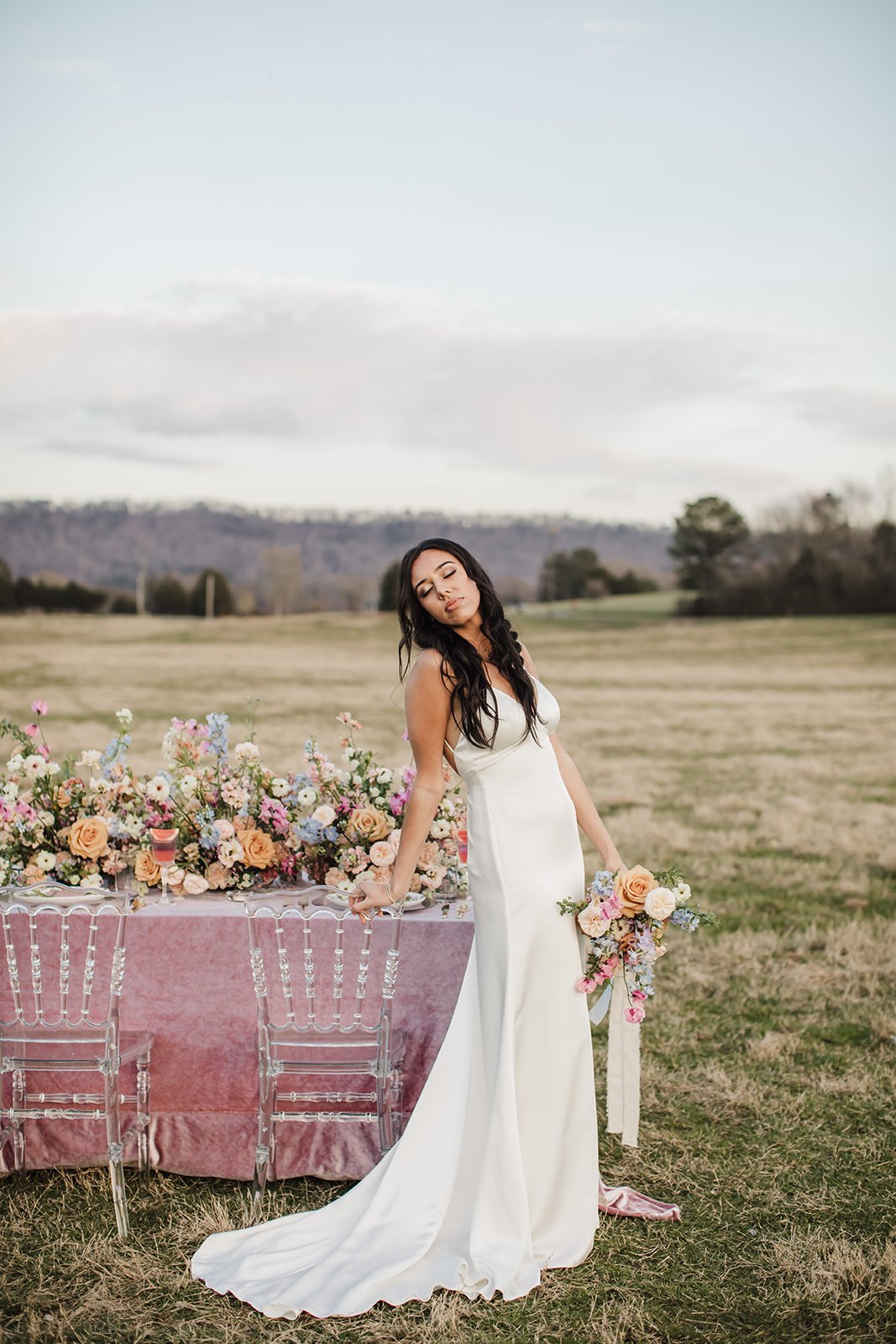 A model holds a pastel bouquet while standing beside a velvet draped table in the middle of a sheep field