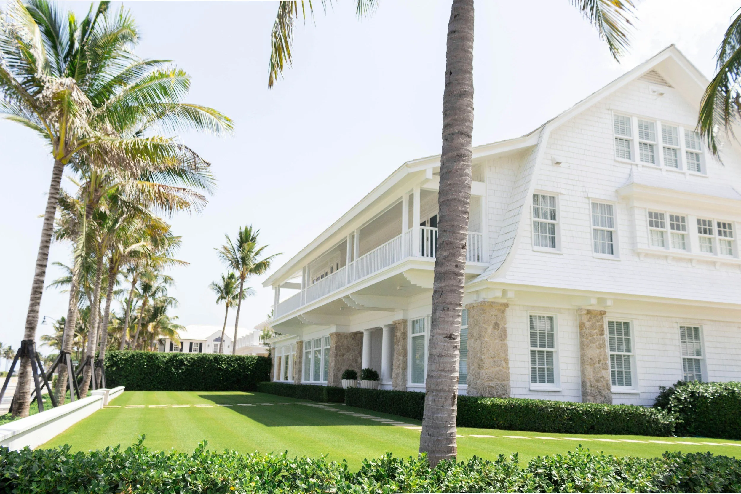 Bright white two-story house with multiple windows, stone accents, and a large covered porch, surrounded by palm trees and a well-maintained lawn