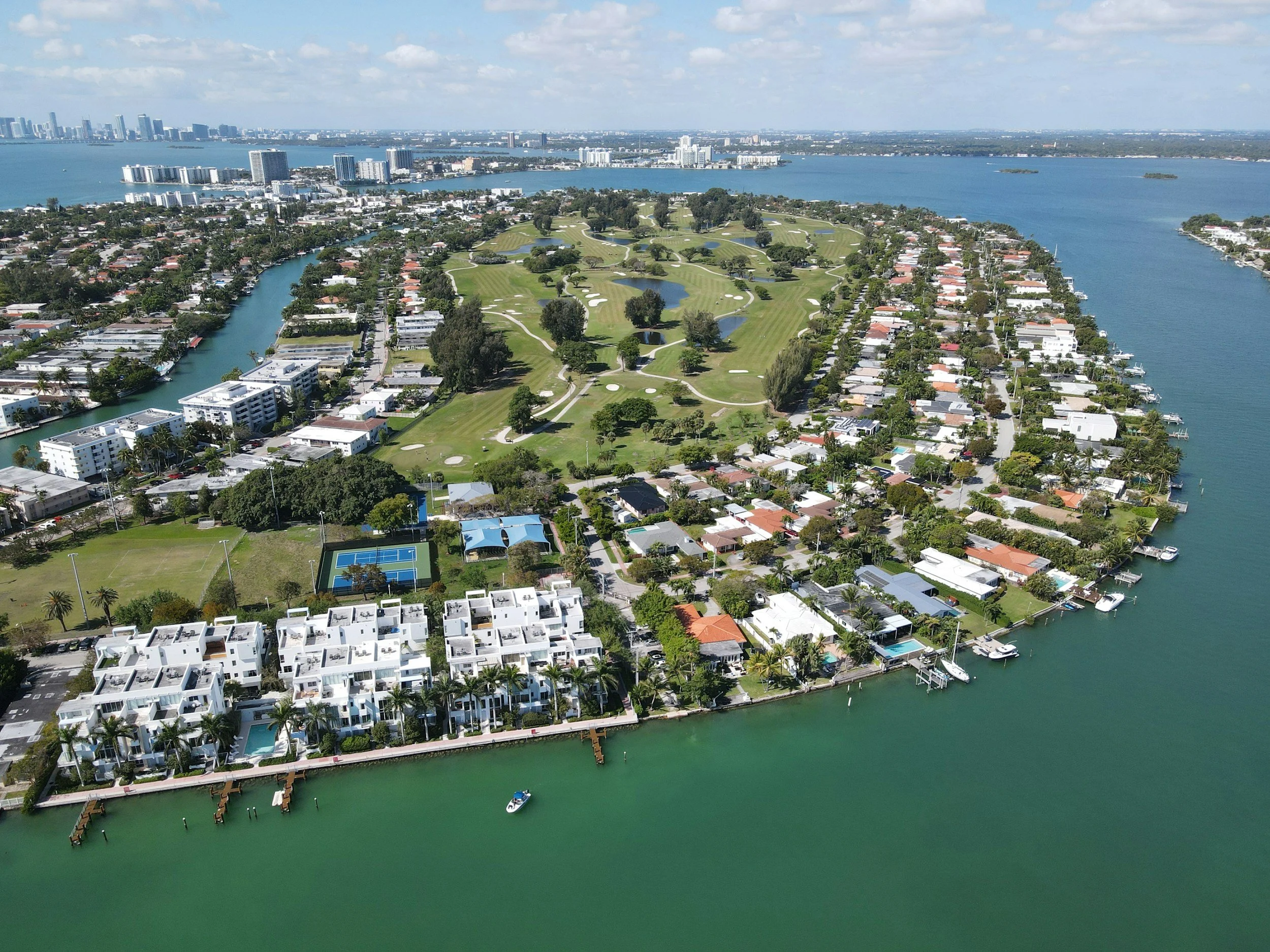 Aerial view of a peninsula with residential houses, a golf course, and a marina along the water; city skyline in the background.