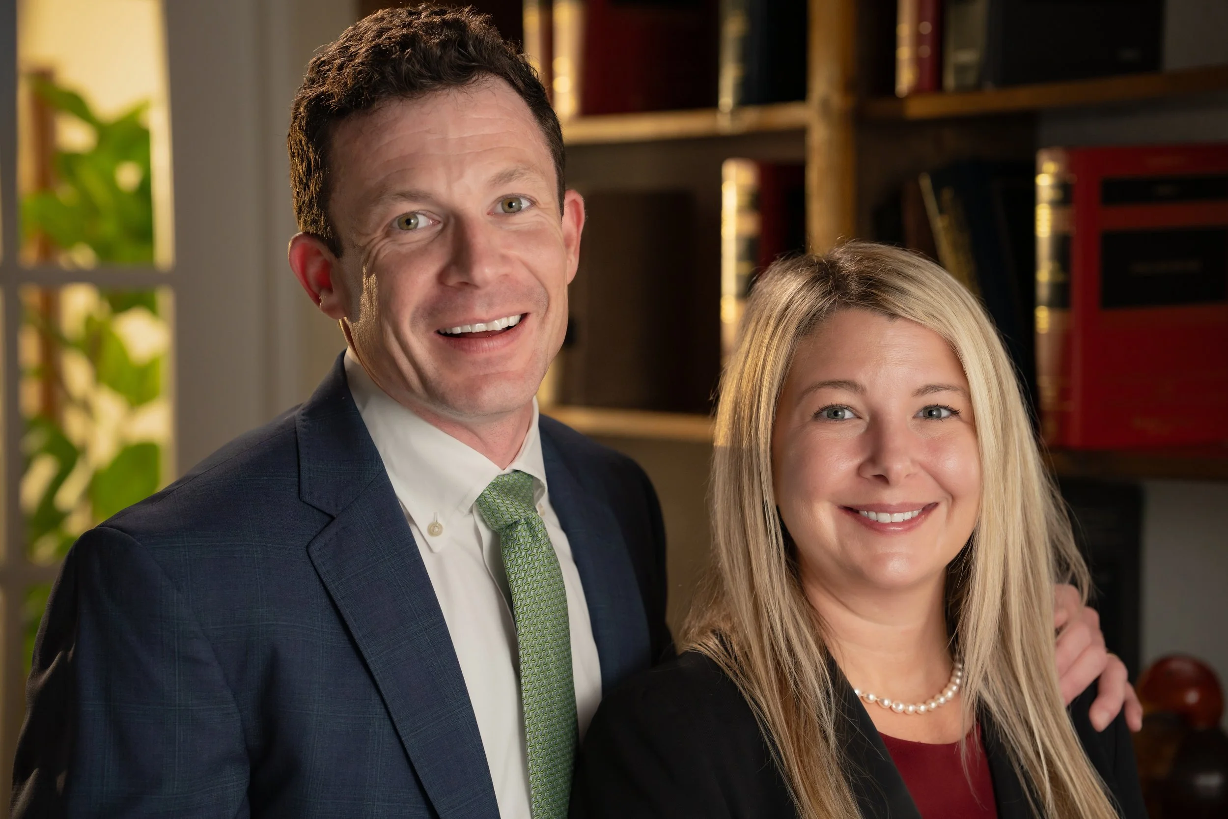 A man in a dark suit and a woman in a black blazer smiling for a photo indoors with a bookshelf in the background.