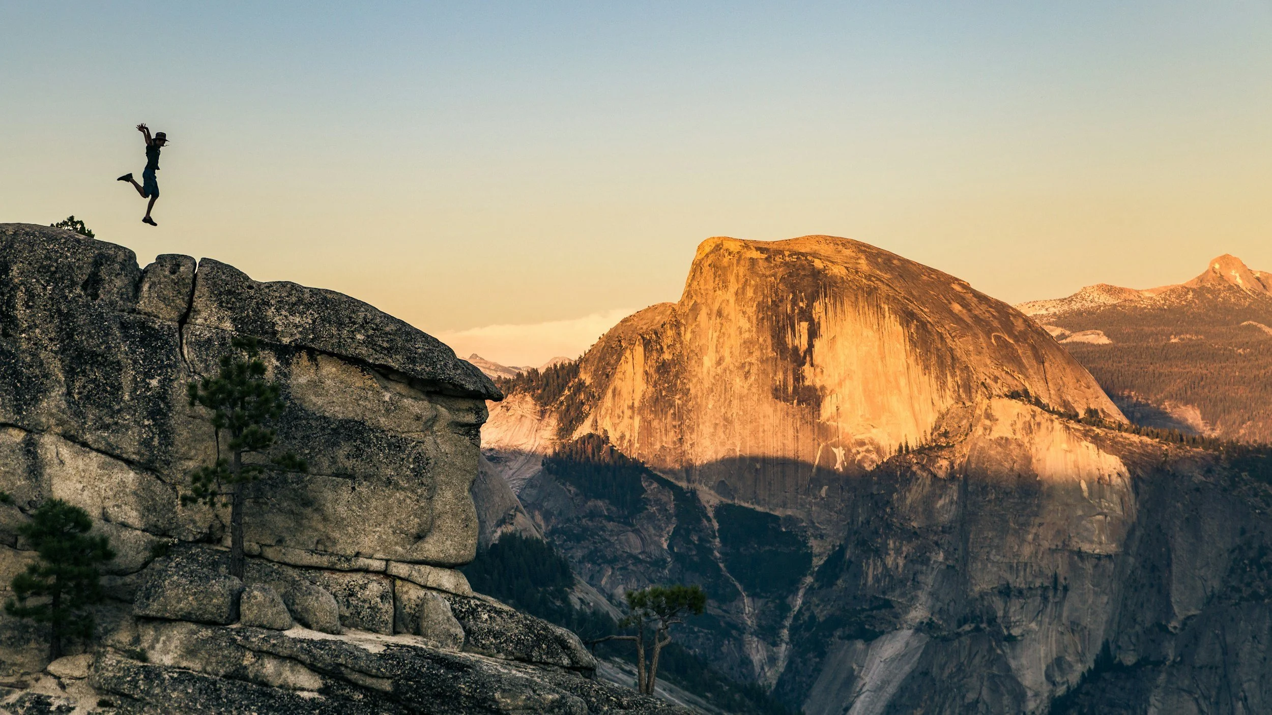 Person jumping off a rocky cliff with a mountain landscape in the background at sunset.