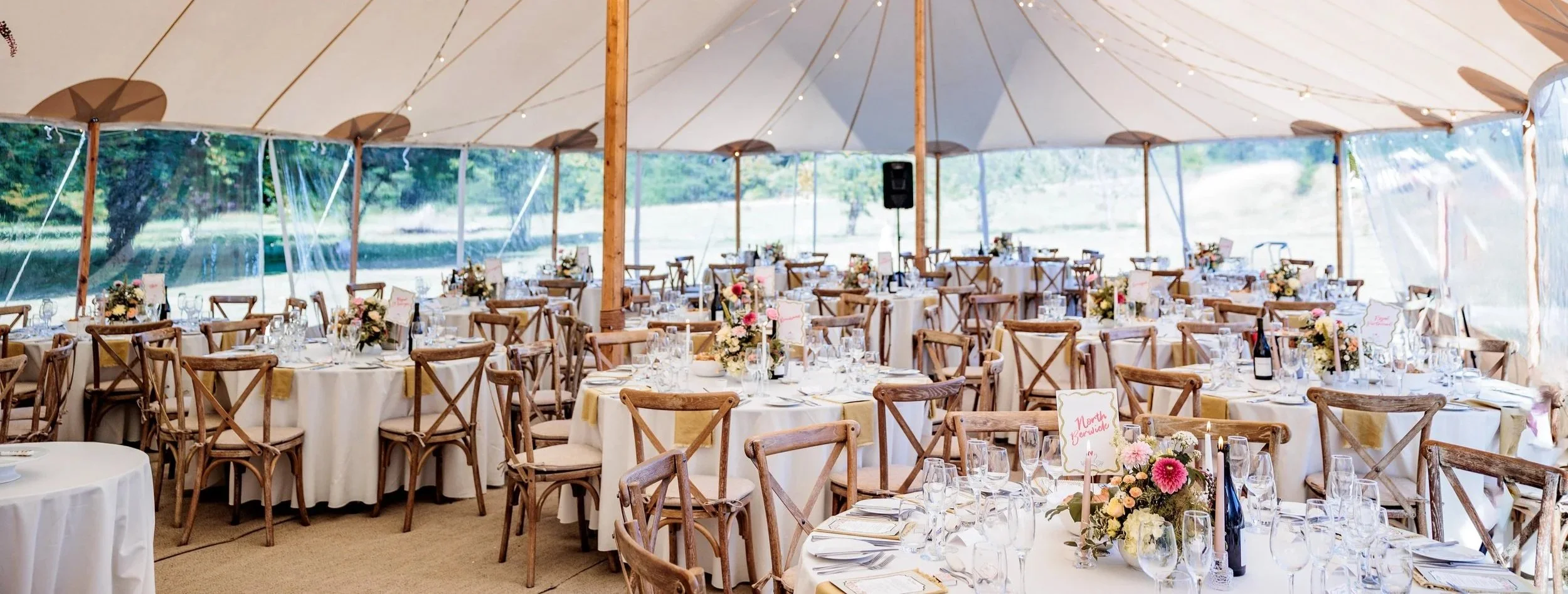 A large outdoor tent set up for a wedding or event with round tables, wooden chairs, floral centerpieces, and table settings under string lights, with a scenic view of trees outside. Sperry tent, isle of wight wedding, planned by Wed & Prosper