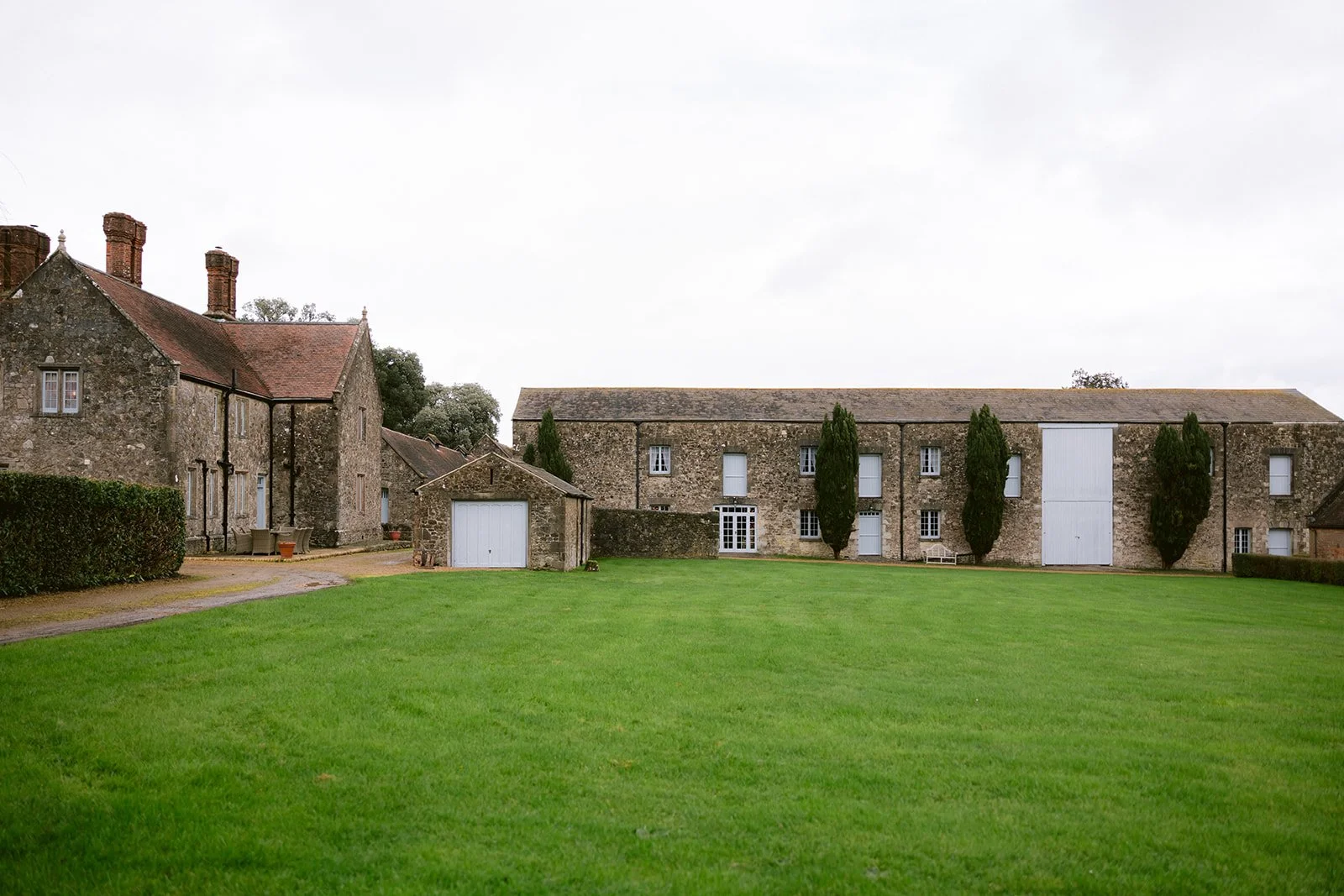 Historic stone buildings with red tile roofs and chimneys, surrounded by a well-maintained green lawn and some tall trees, under a cloudy sky. Isle of wight wedding venue