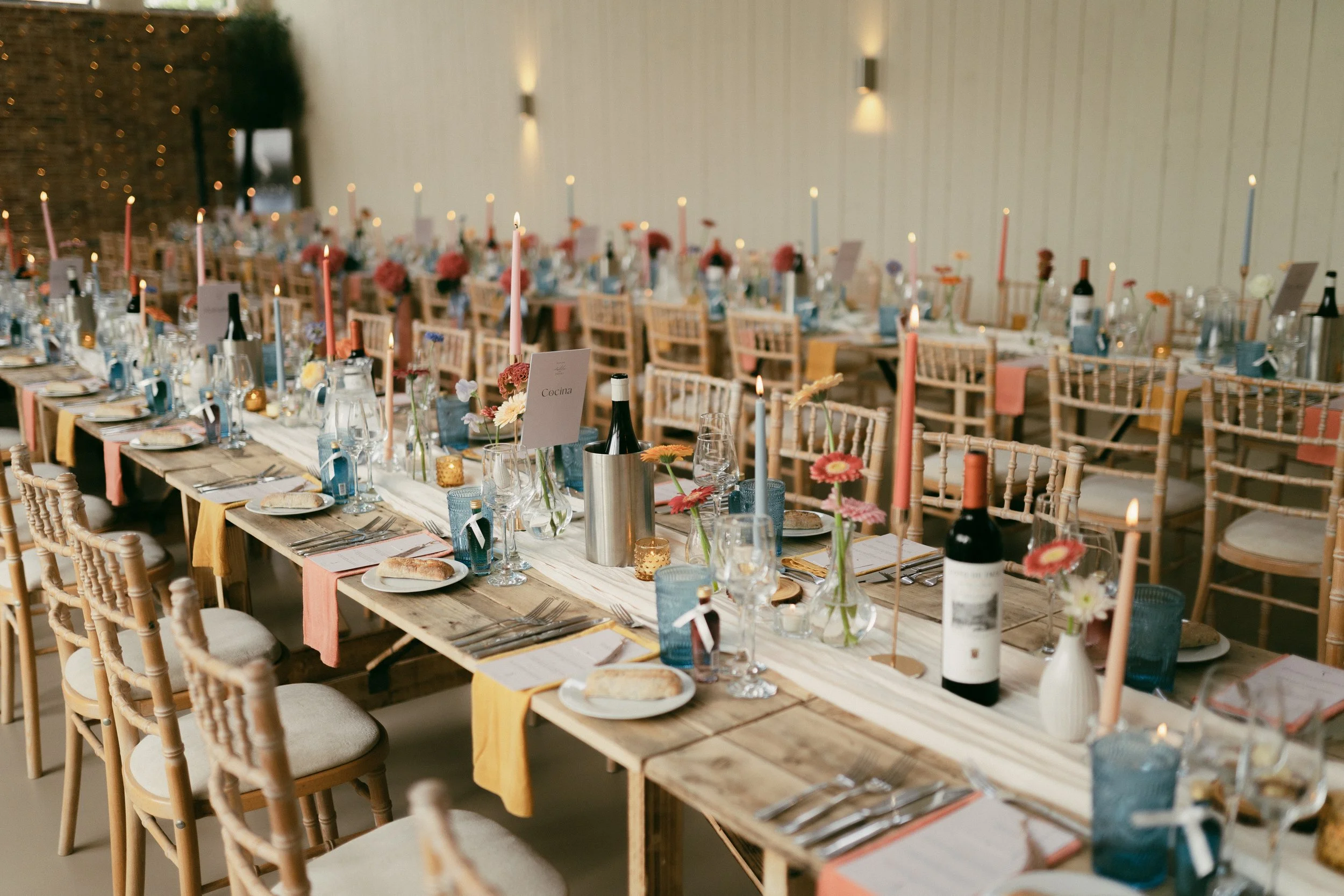 Elegant banquet table setup with wooden tables, pastel-colored table runners, floral centerpieces, pink and blue taper candles, wine bottles, and neatly arranged glassware and silverware in a decorated reception hall.