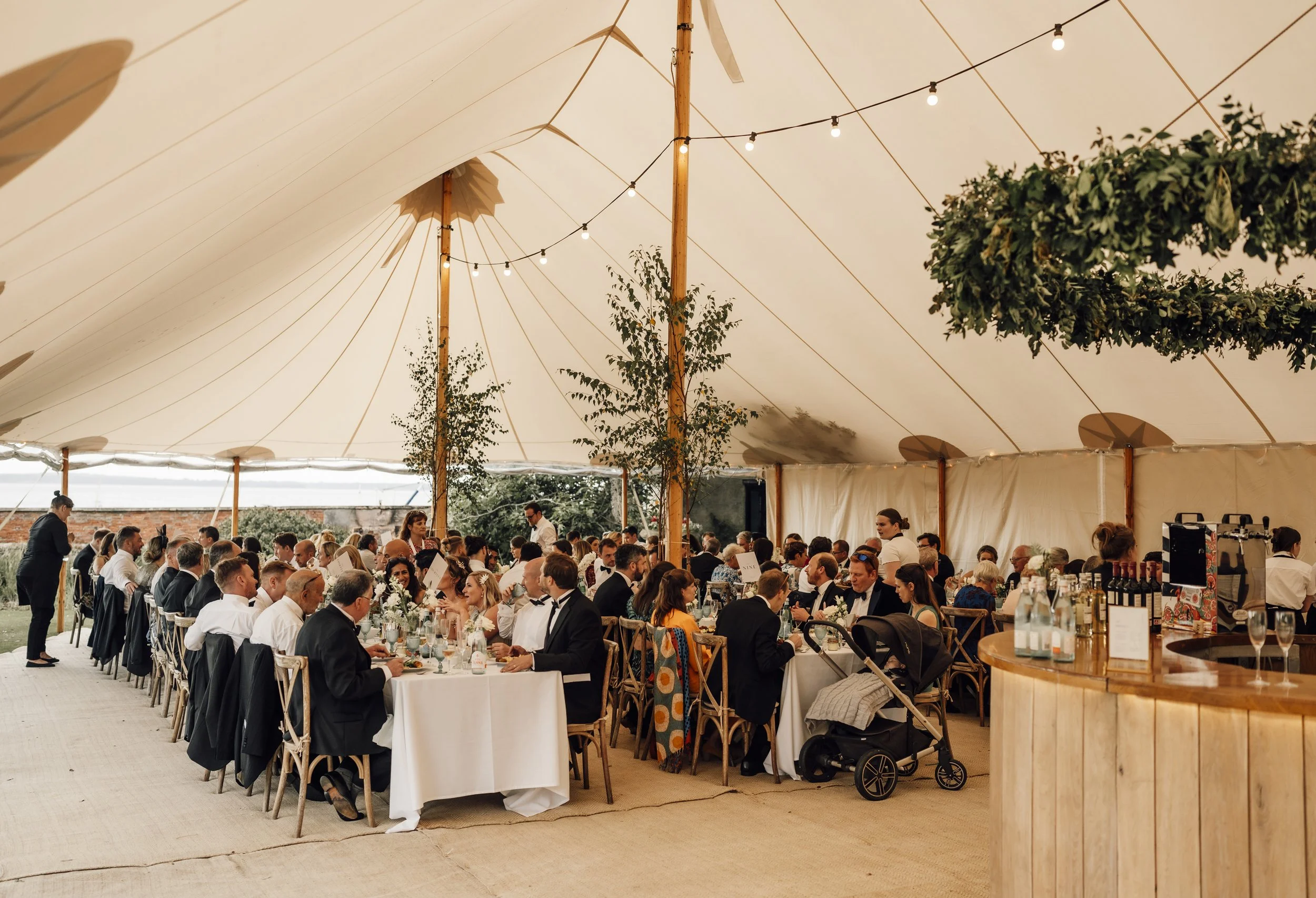 Wedding reception inside a large tent with many guests seated at decorated tables, string lights hanging from the ceiling, and greenery outside visible through the open sides. Floral installations by wed & prosper