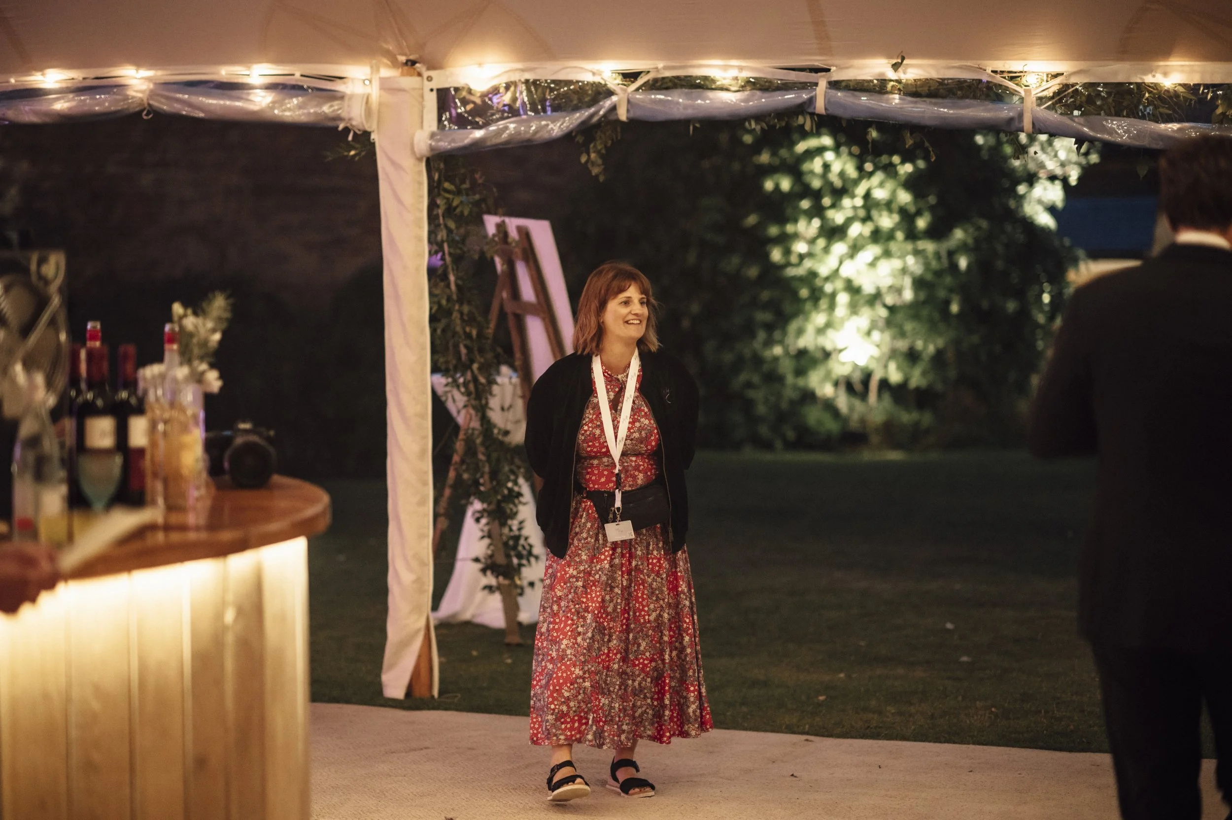 A woman with short red hair, wearing a red patterned dress with a black jacket and sandals, stands smiling at an outdoor evening event under a decorated tent. Carrie award winning wedding planner uk
