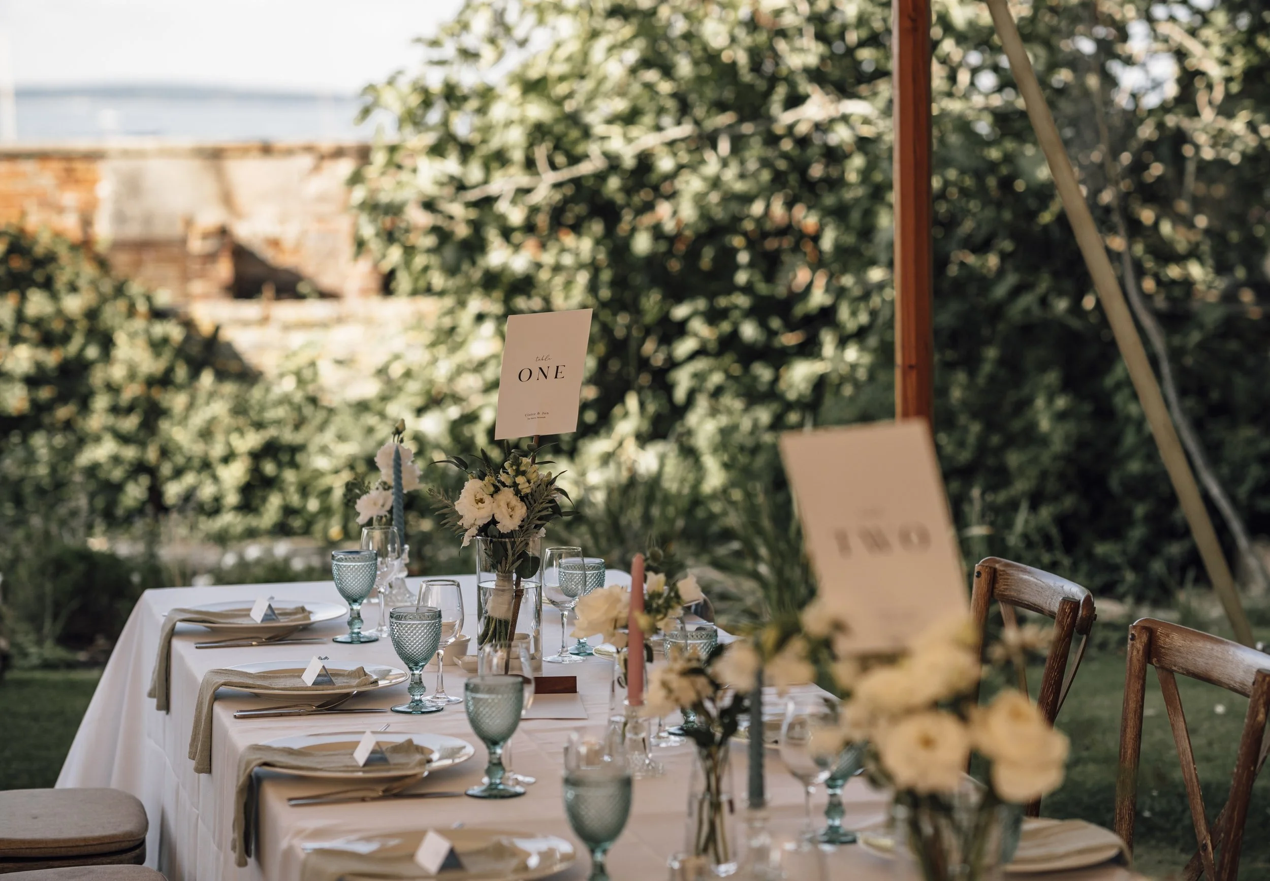 Elegant outdoor table set for a celebration, decorated with white flowers, candles, and glassware, labeled with table signs marked 'ONE' and 'TWO', surrounded by wooden chairs, with greenery and a brick wall in the background.