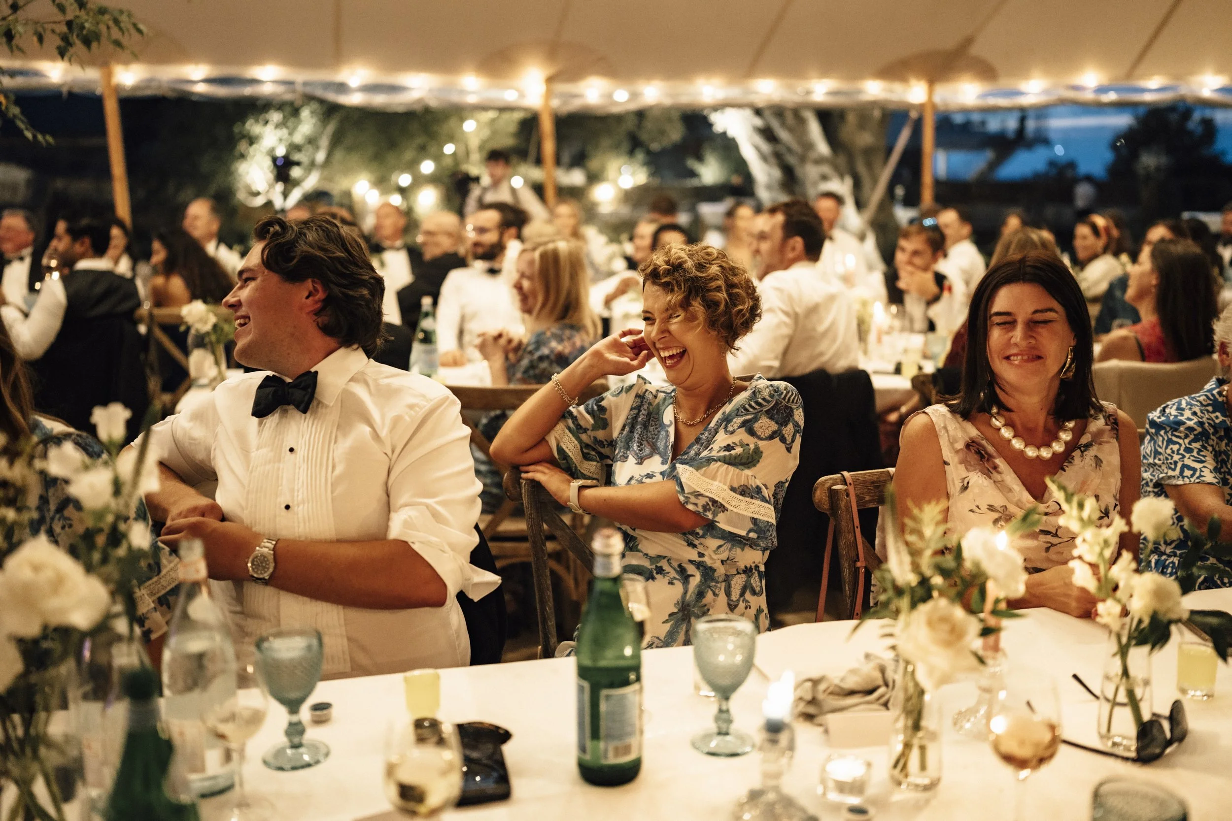 People sitting at decorated tables, some laughing and smiling, at a formal evening event or celebration under a tent with string lights.