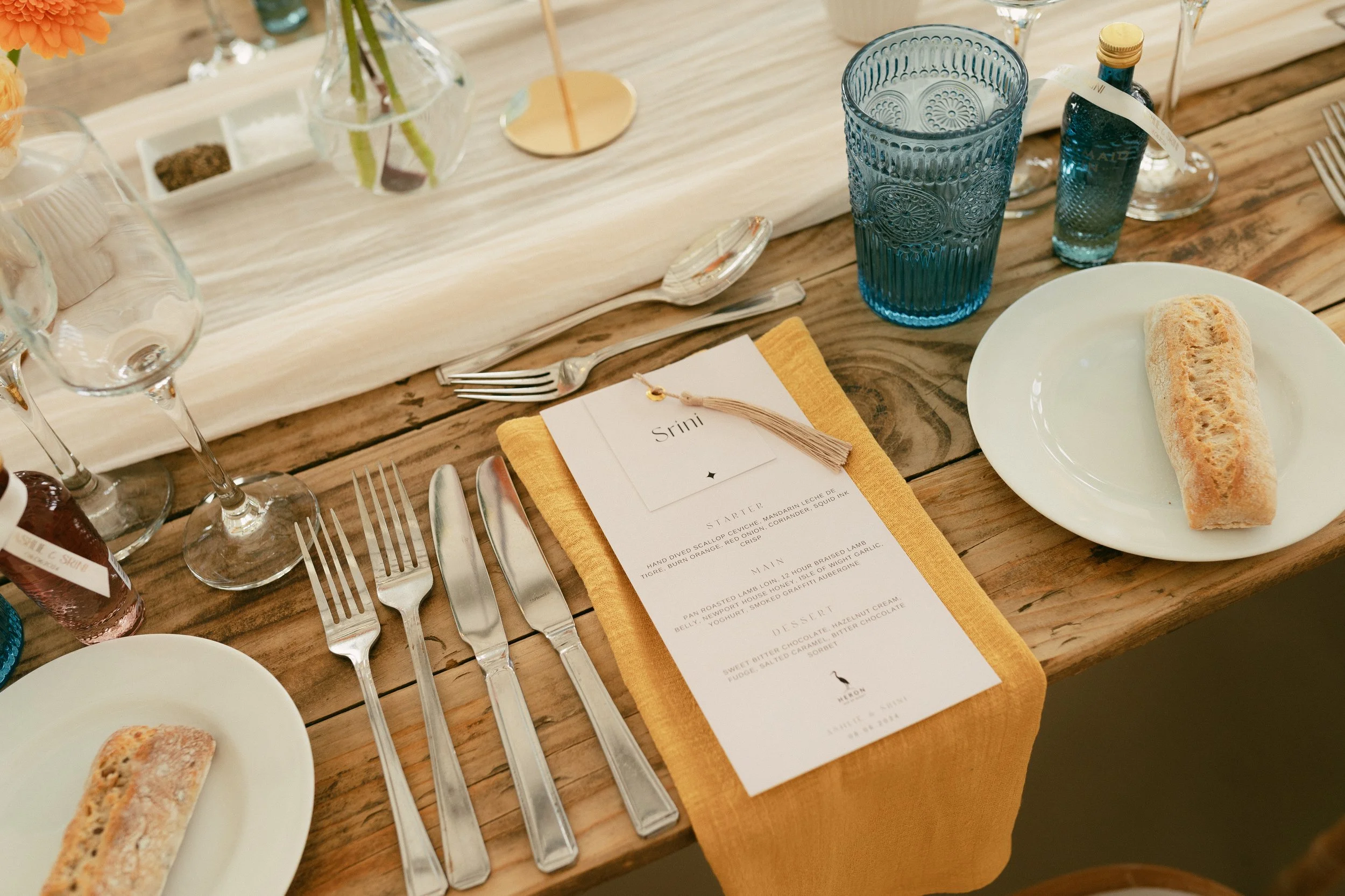 A rustic wooden dining table set with white plates, each holding a baguette, silverware, a yellow napkin, a blue glass, a small bottle of blue liquor, and a printed menu with a tassel on top. There is also a centerpiece with a glass vase containing flowers