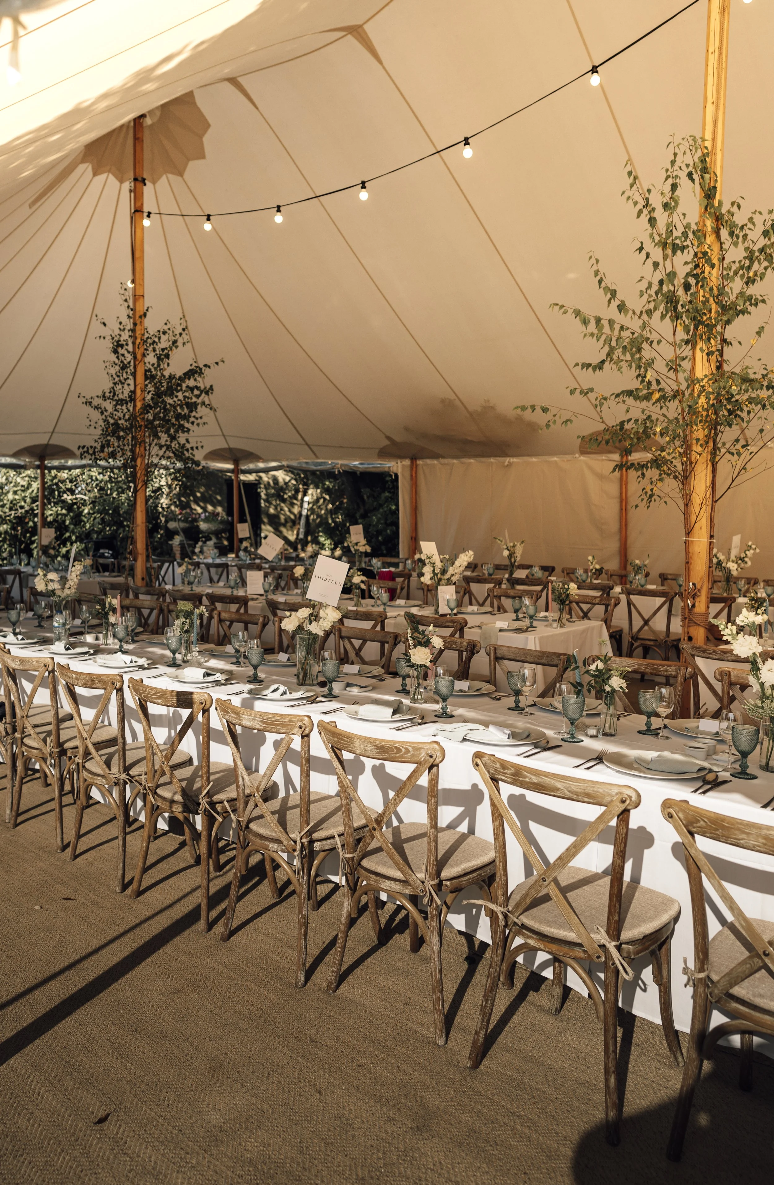 A long banquet table set for an event inside a large white tent, decorated with flower arrangements, green glasses, and place settings, with string lights hanging from the ceiling.