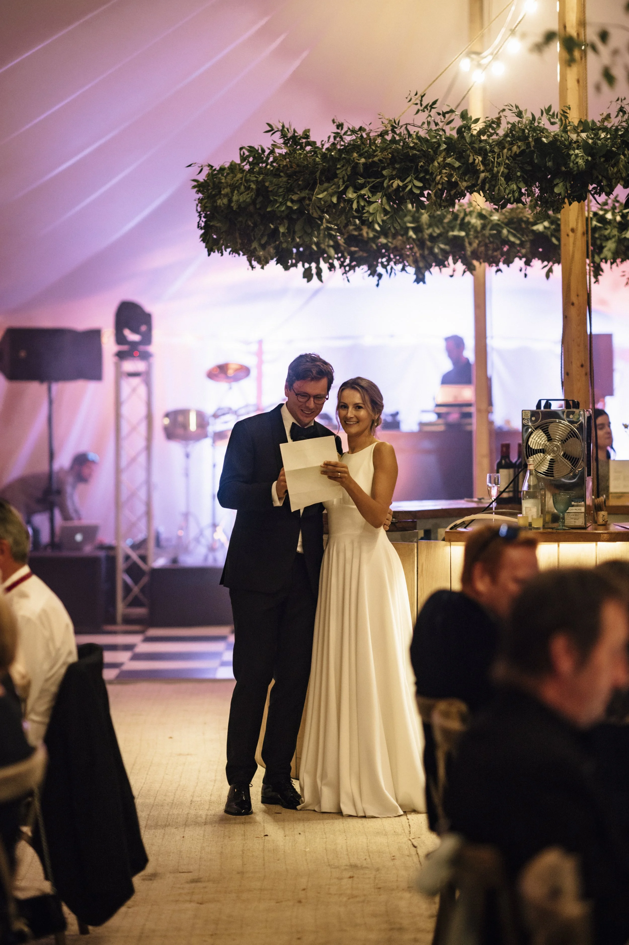 A bride and groom sharing a moment together at their wedding reception, with the bride in a white gown and the groom in a black tuxedo, standing under a decorated canopy with string lights in the background.