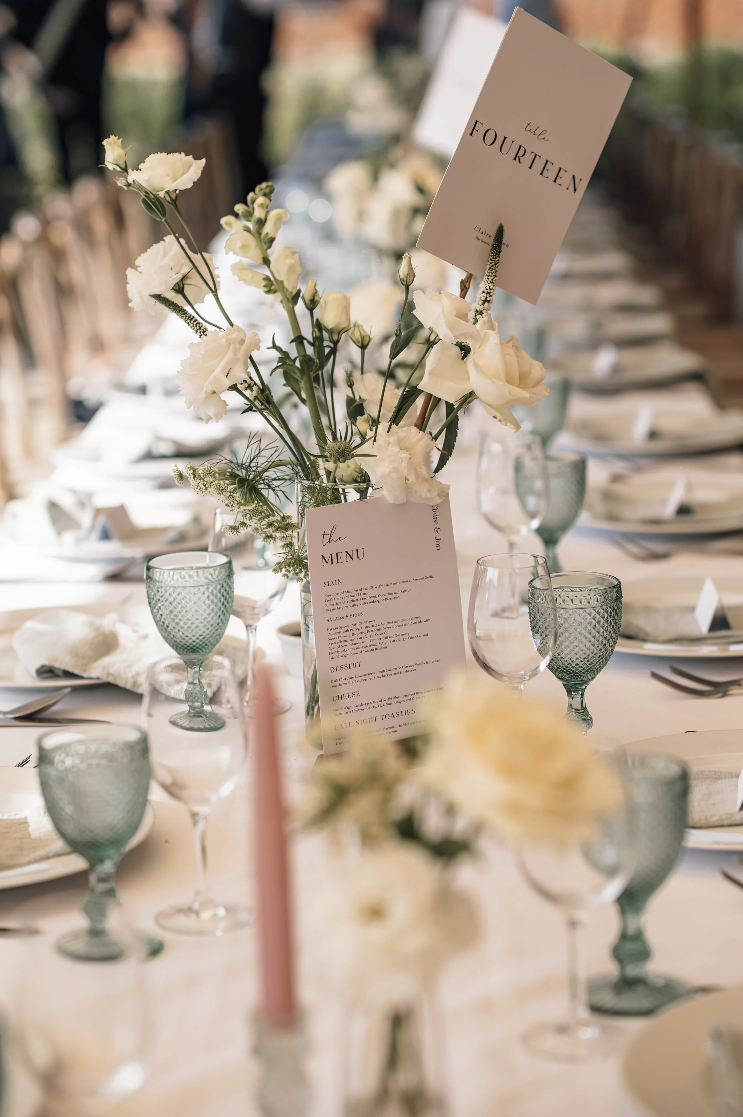 Decorated wedding reception table with white flowers, pink candles, wine glasses, and a menu card in a floral centerpiece, with a seating chart displaying table number fourteen.