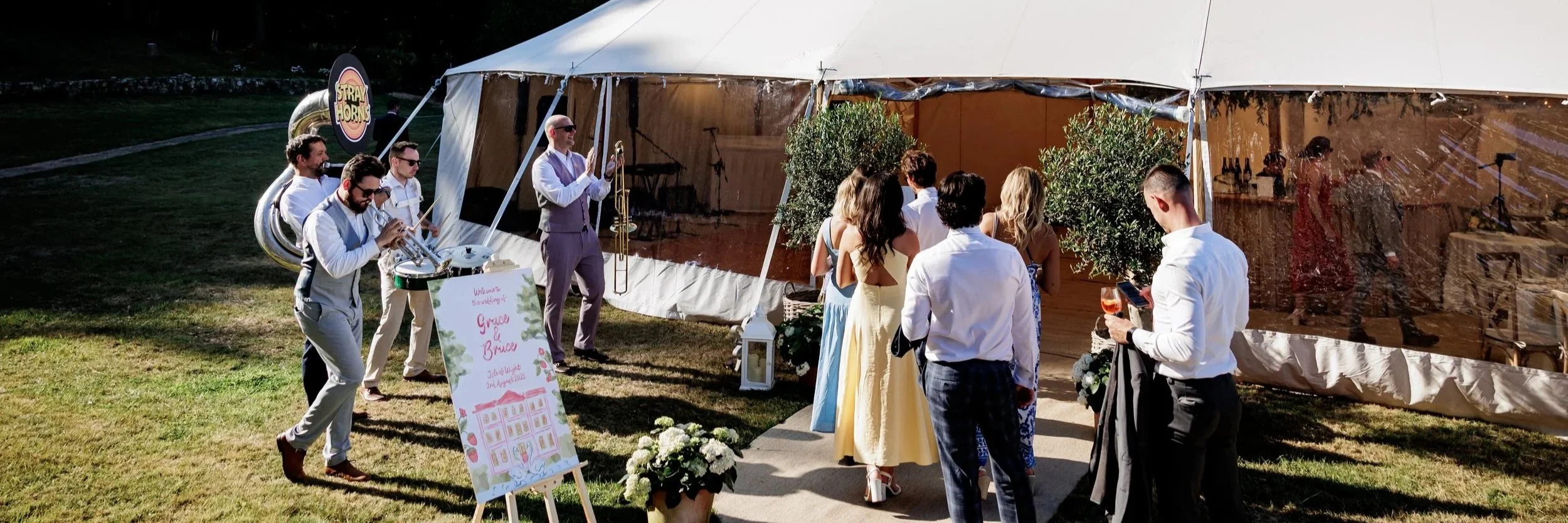 Guests at an outdoor wedding reception, standing near a tent with a band playing music.