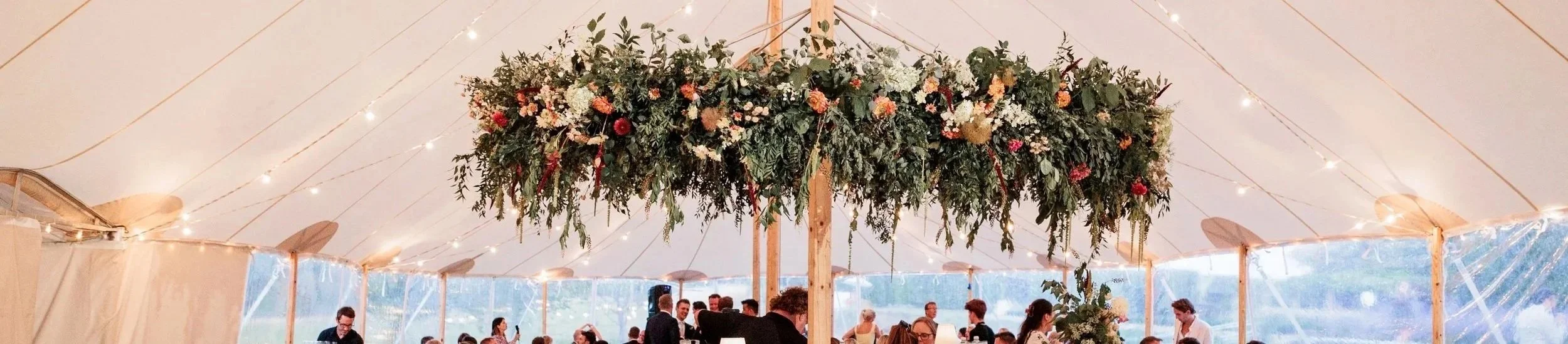 Wedding reception under a large tent with a floral arrangement hanging from the ceiling and string lights, with guests seated at tables. large floral hoop, Sperry tent, Isle of wight wedding