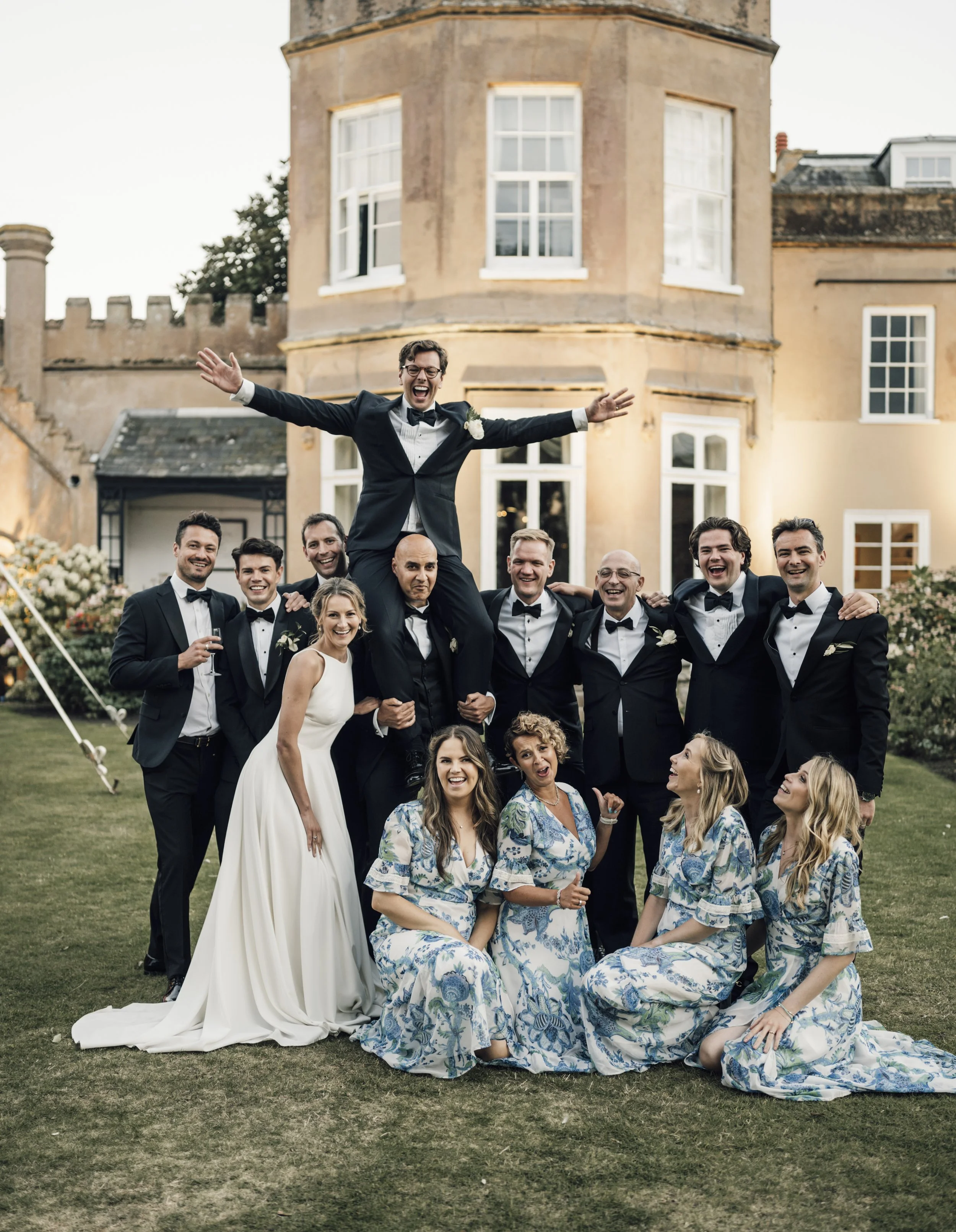 A wedding party celebrating outdoors in front of a large historic building, with a man being lifted into the air and others smiling and posing for the photo.