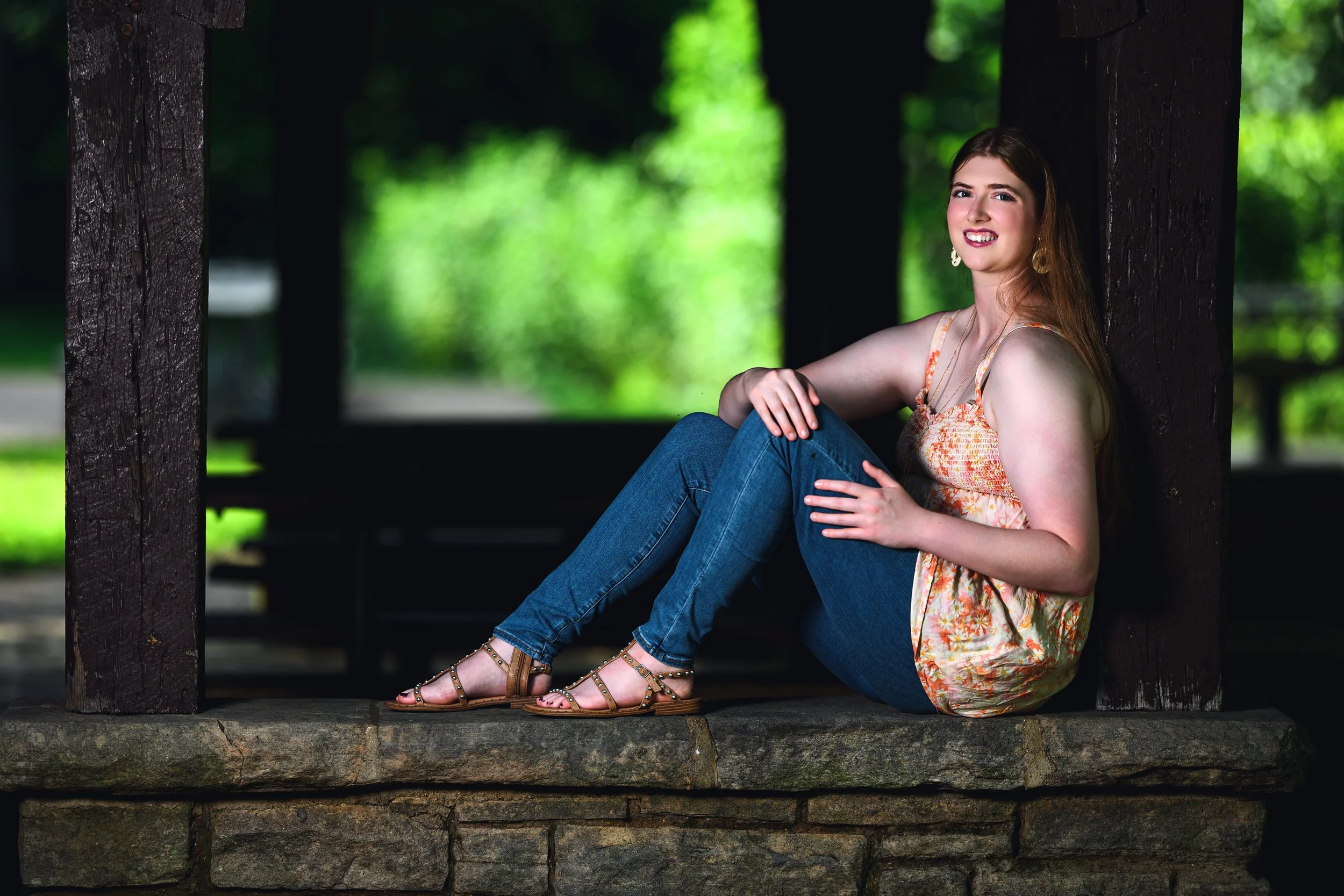A young woman with long red hair sitting on a stone ledge in a shaded park, wearing a floral top, jeans, and sandals, smiling at the camera.