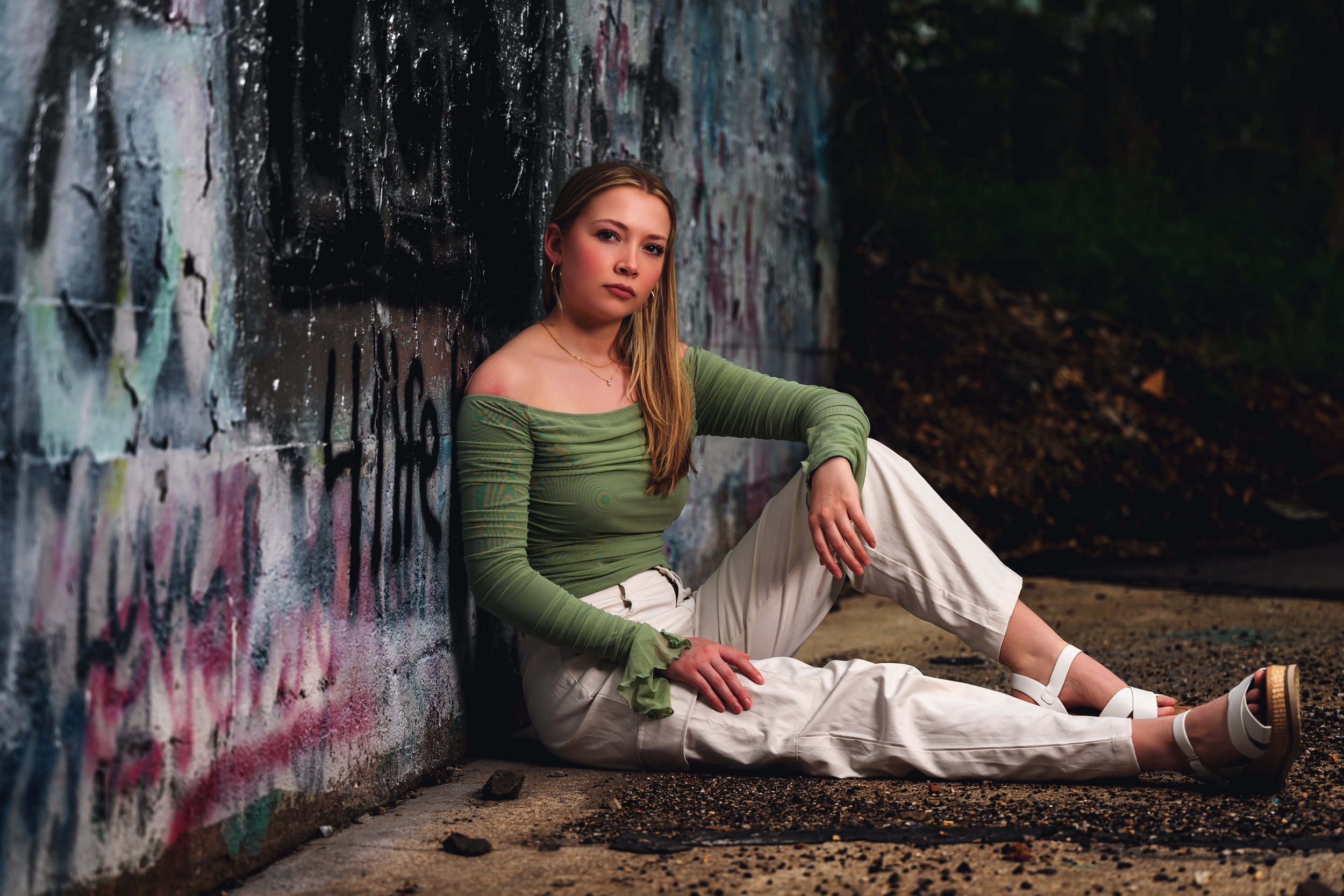 A person in a green off-shoulder top and white pants sitting on the ground against a graffiti-covered wall.