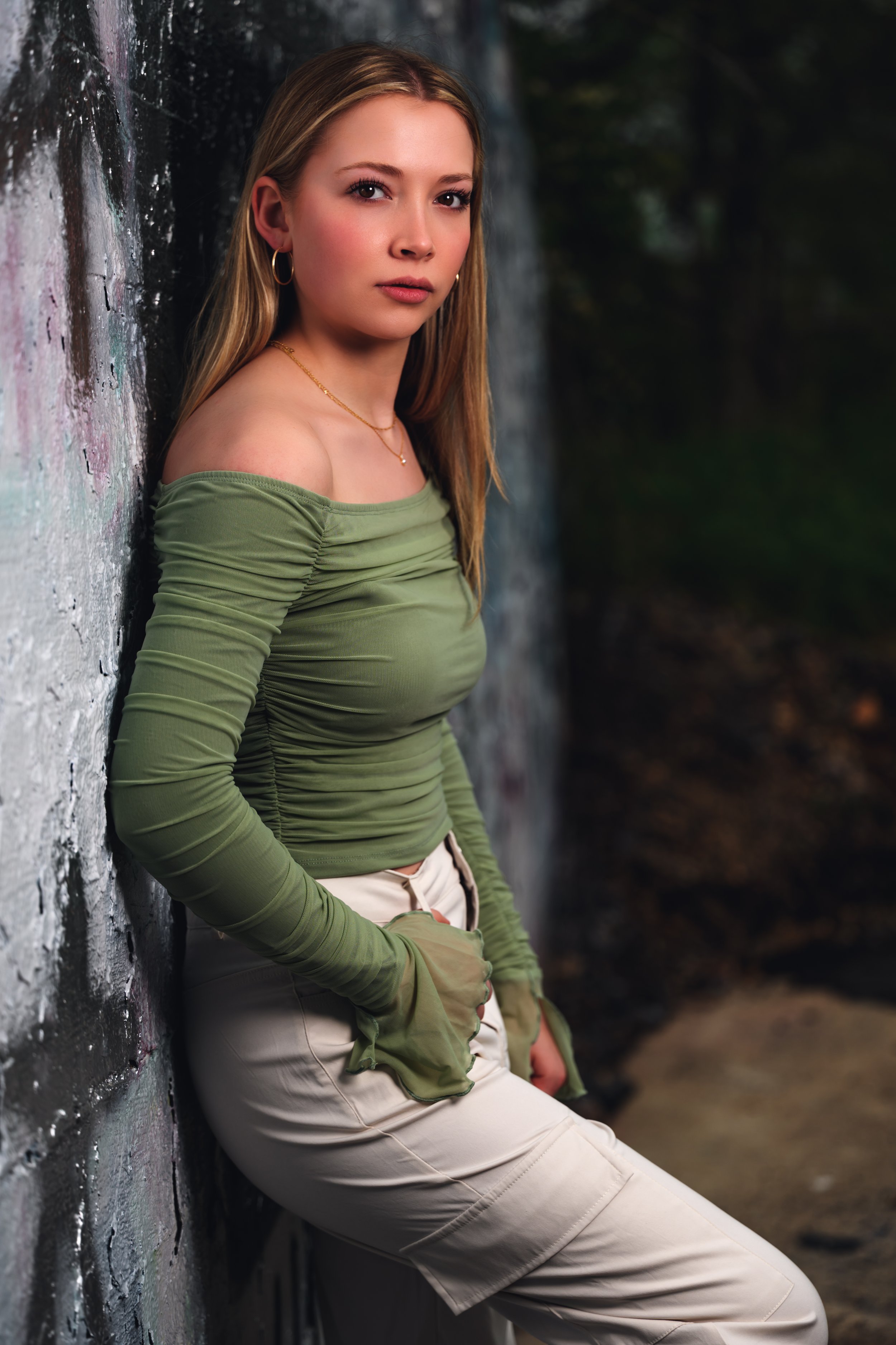 A woman in a green off-shoulder top and beige pants leaning against a textured wall outdoors.