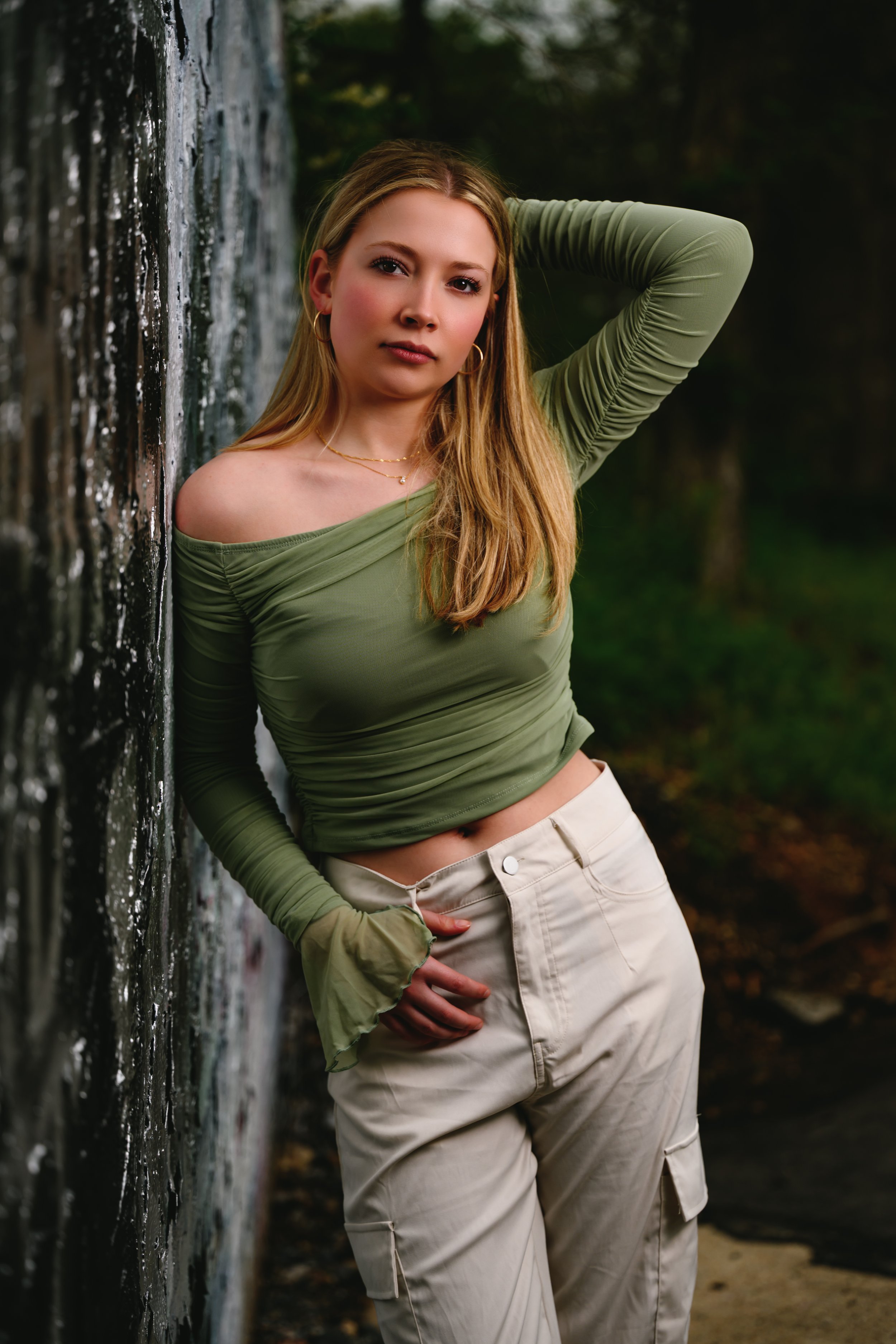 Person wearing a green off-shoulder top and light-colored cargo pants leaning against a graffiti-covered wall, with trees in the background.