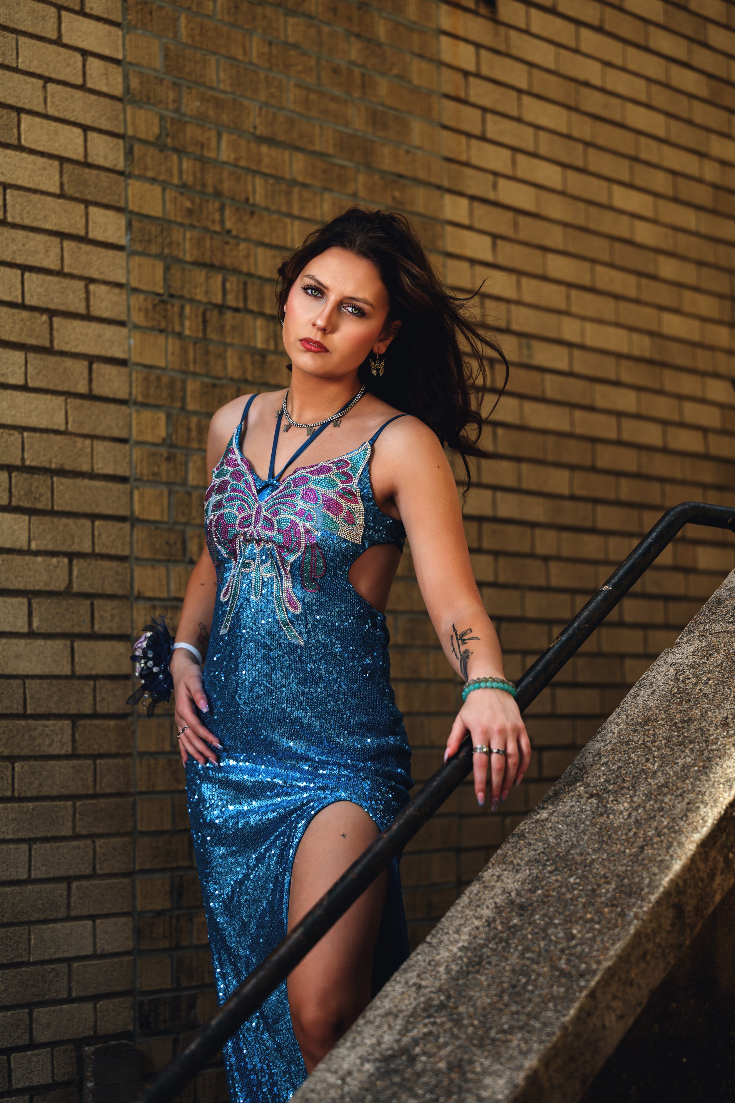Woman in a blue sequined dress with butterfly design leaning on a staircase railing against a brick wall.