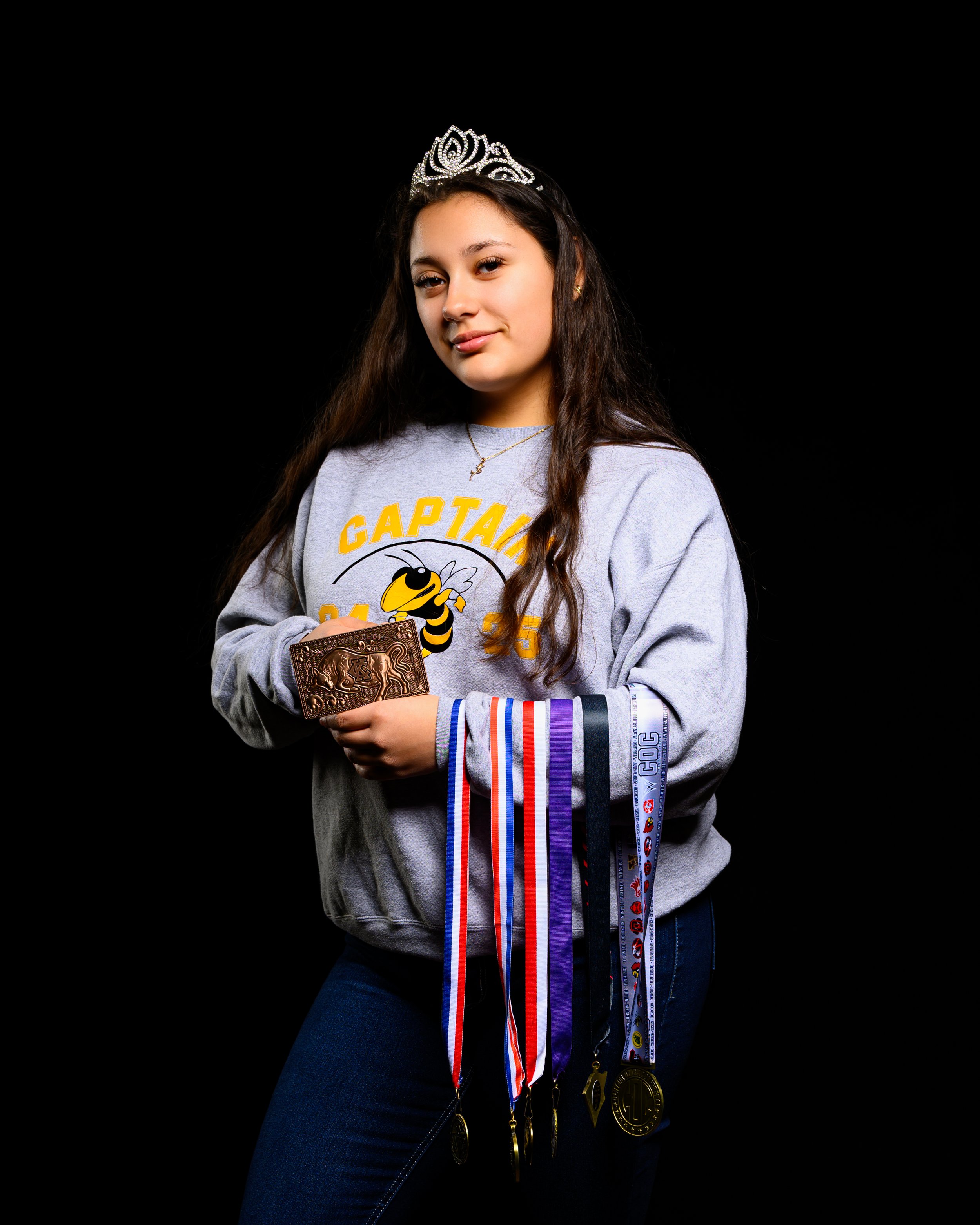 Young woman wearing a tiara and a sweatshirt with a bee logo, holding medals and a trophy, set against a black background.