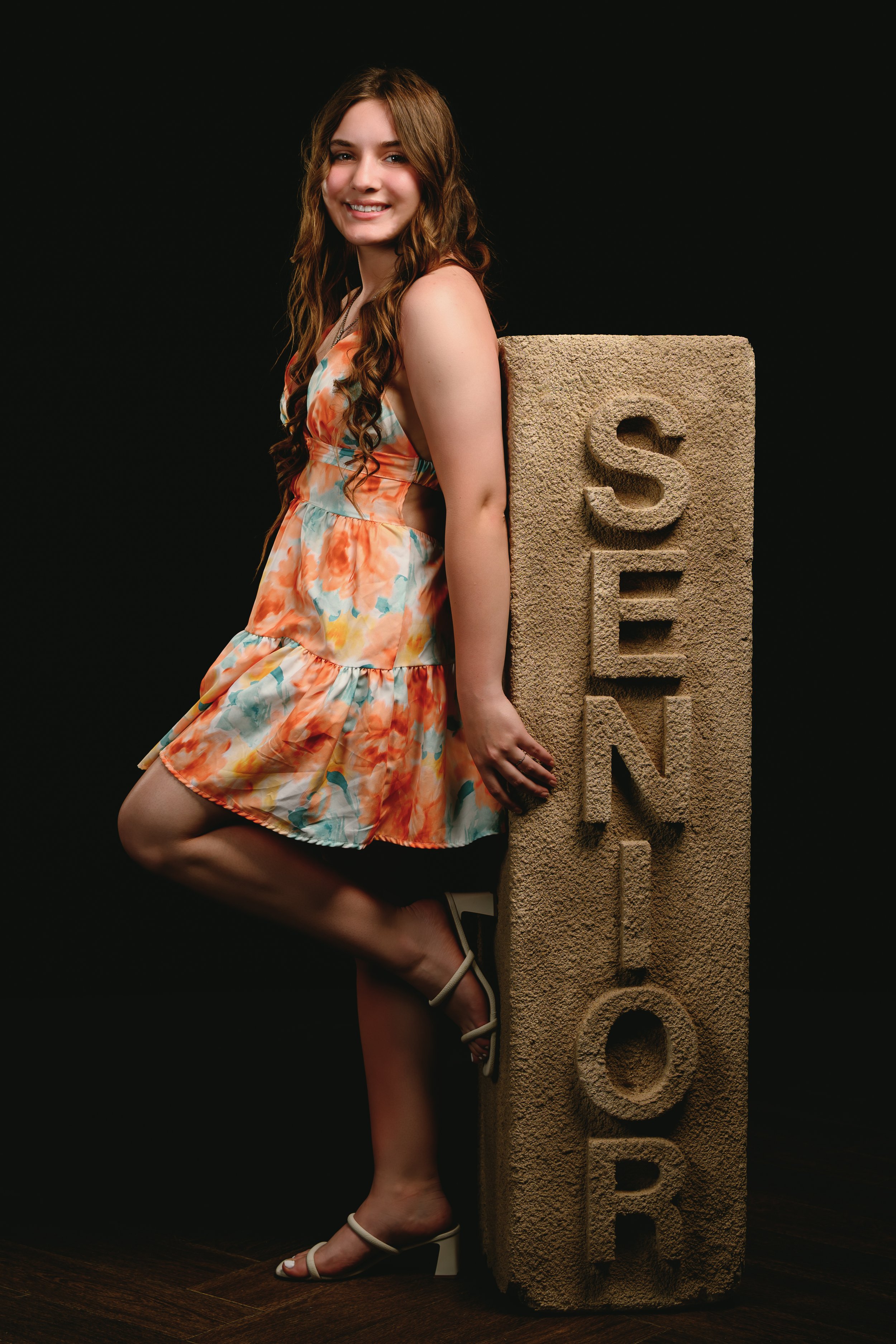 A person in a colorful floral dress leans against a stone sculpture with the word 'SENIOR' carved into it.