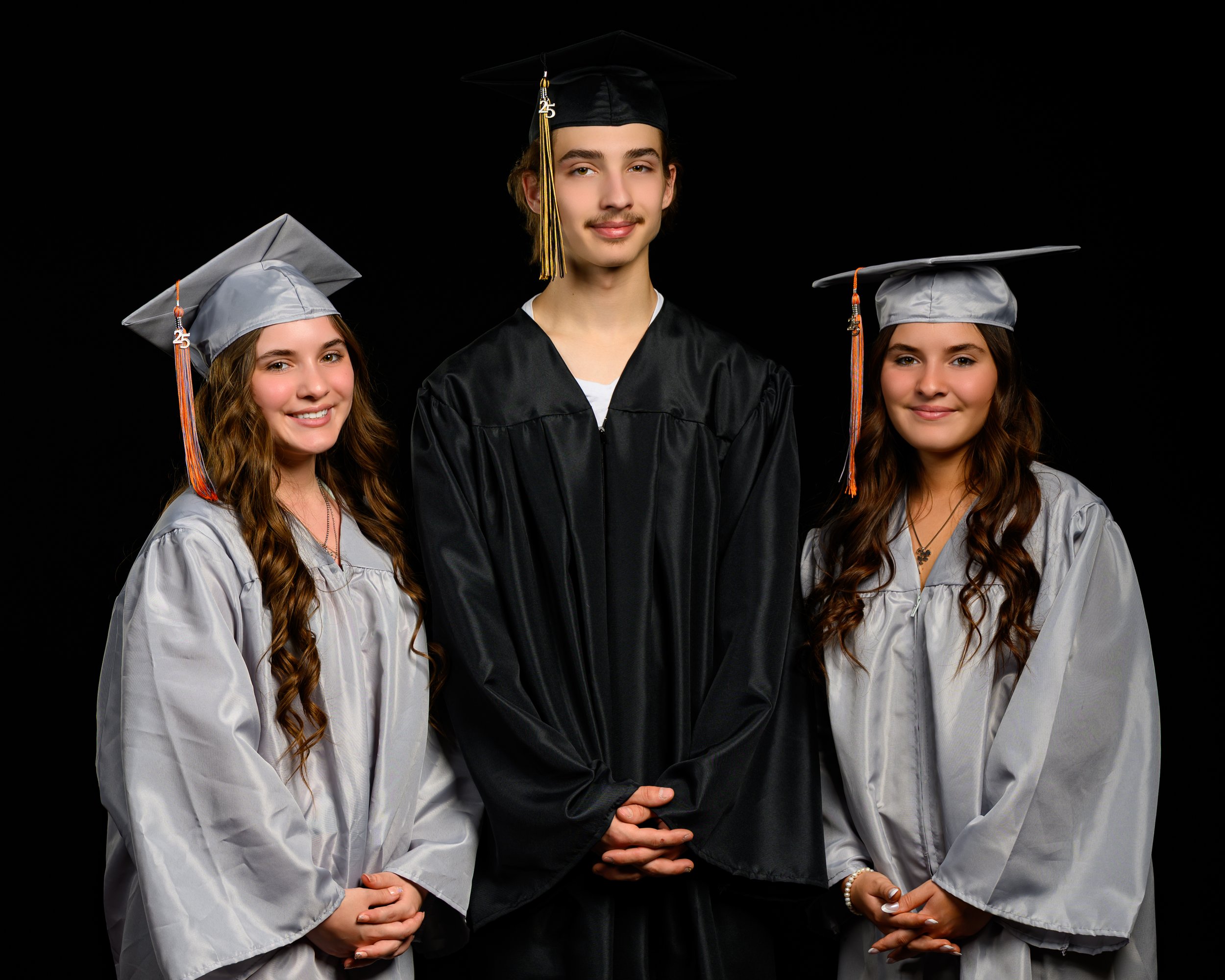 Three graduates posing in caps and gowns, two in silver and one in black, against a black background.