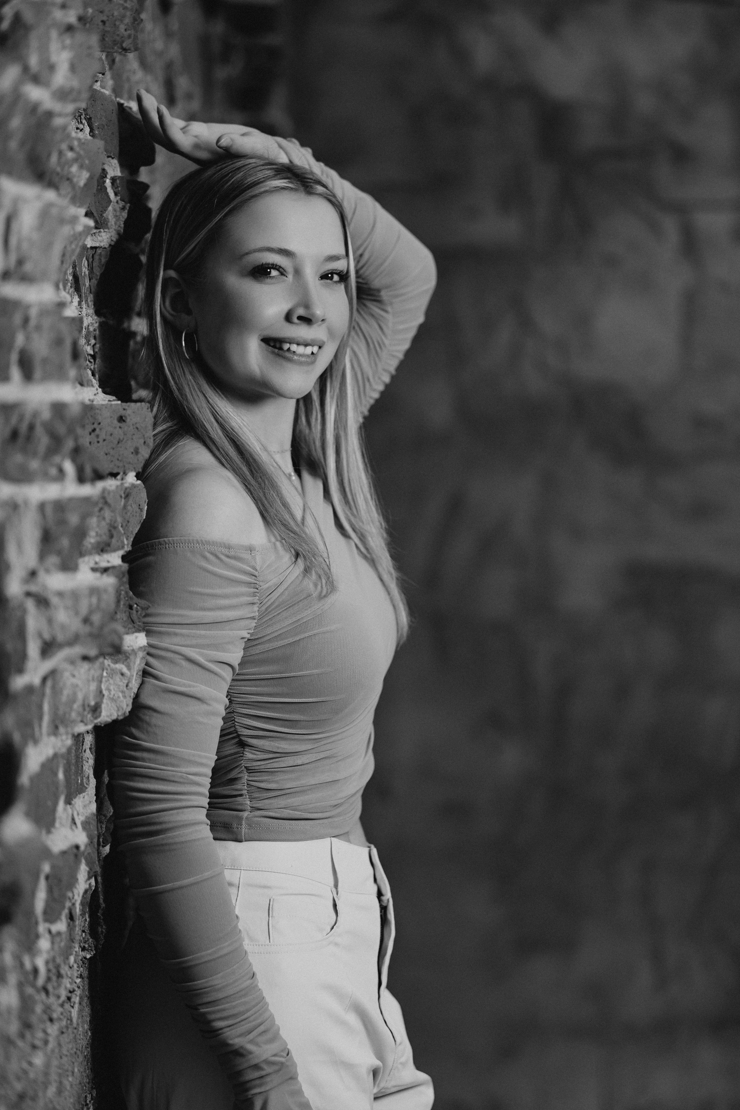 Black and white portrait of a smiling woman leaning against a brick wall, with one hand touching her head.