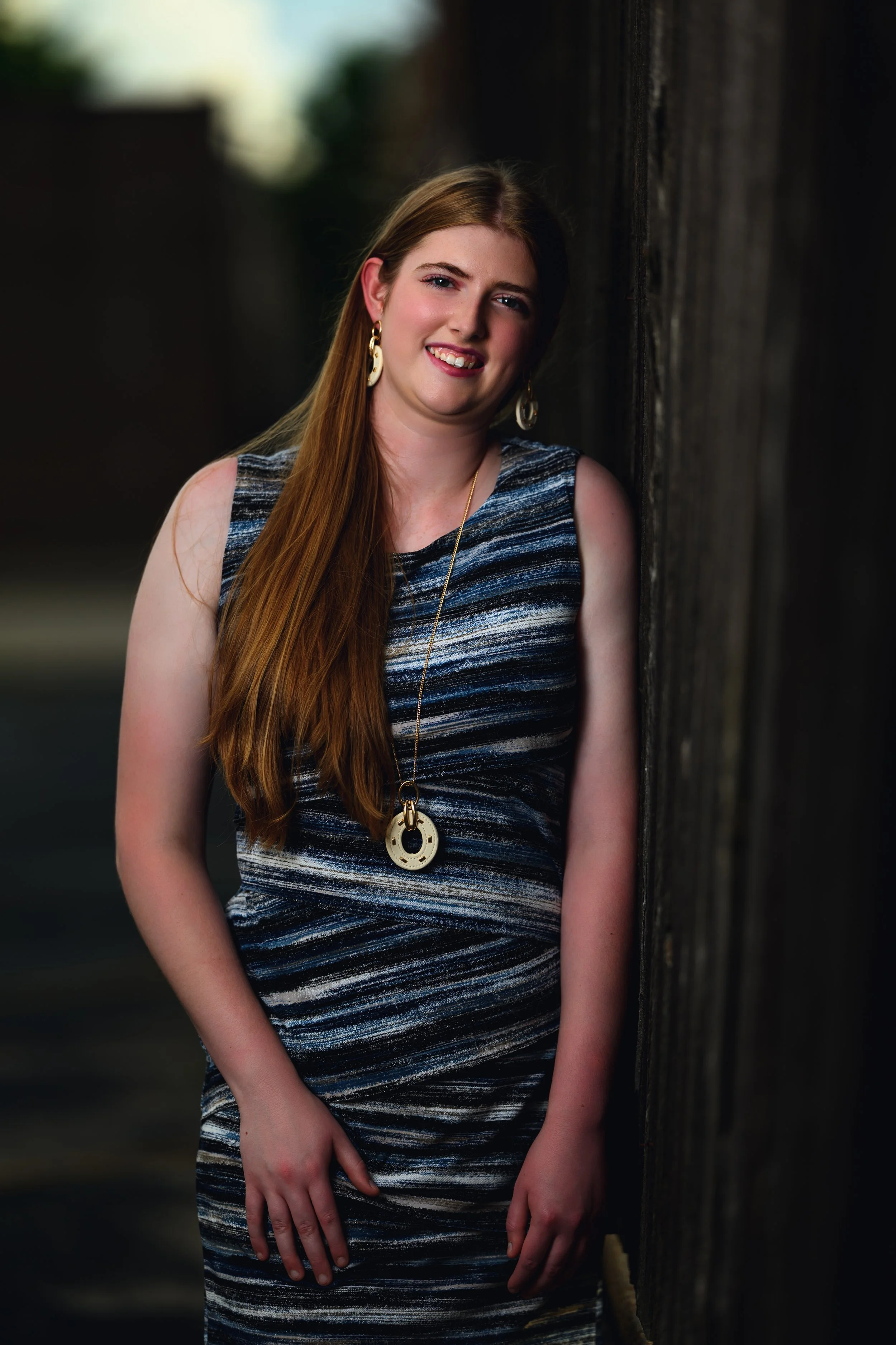 A young woman with long red hair, wearing a sleeveless blue and black patterned dress, gold earrings, and a long necklace, leaning against a dark wooden wall outdoors at sunset.