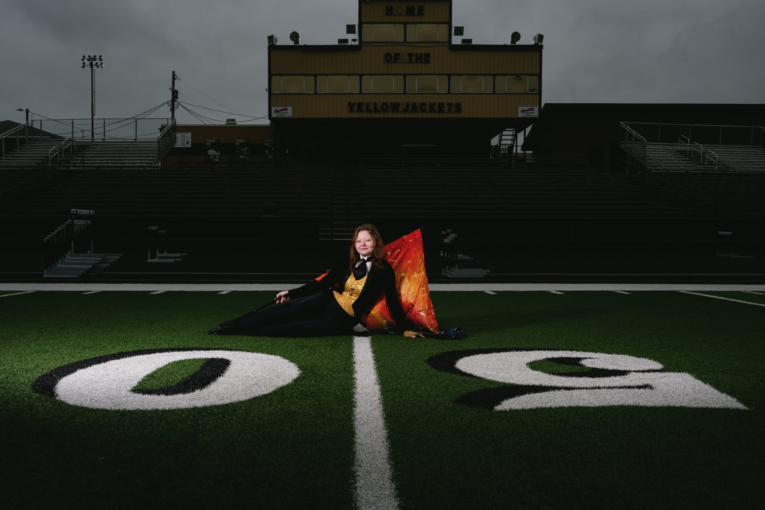 Person in marching band uniform with colorful flag, sitting on a football field's 50-yard line, with stadium bleachers in the background.