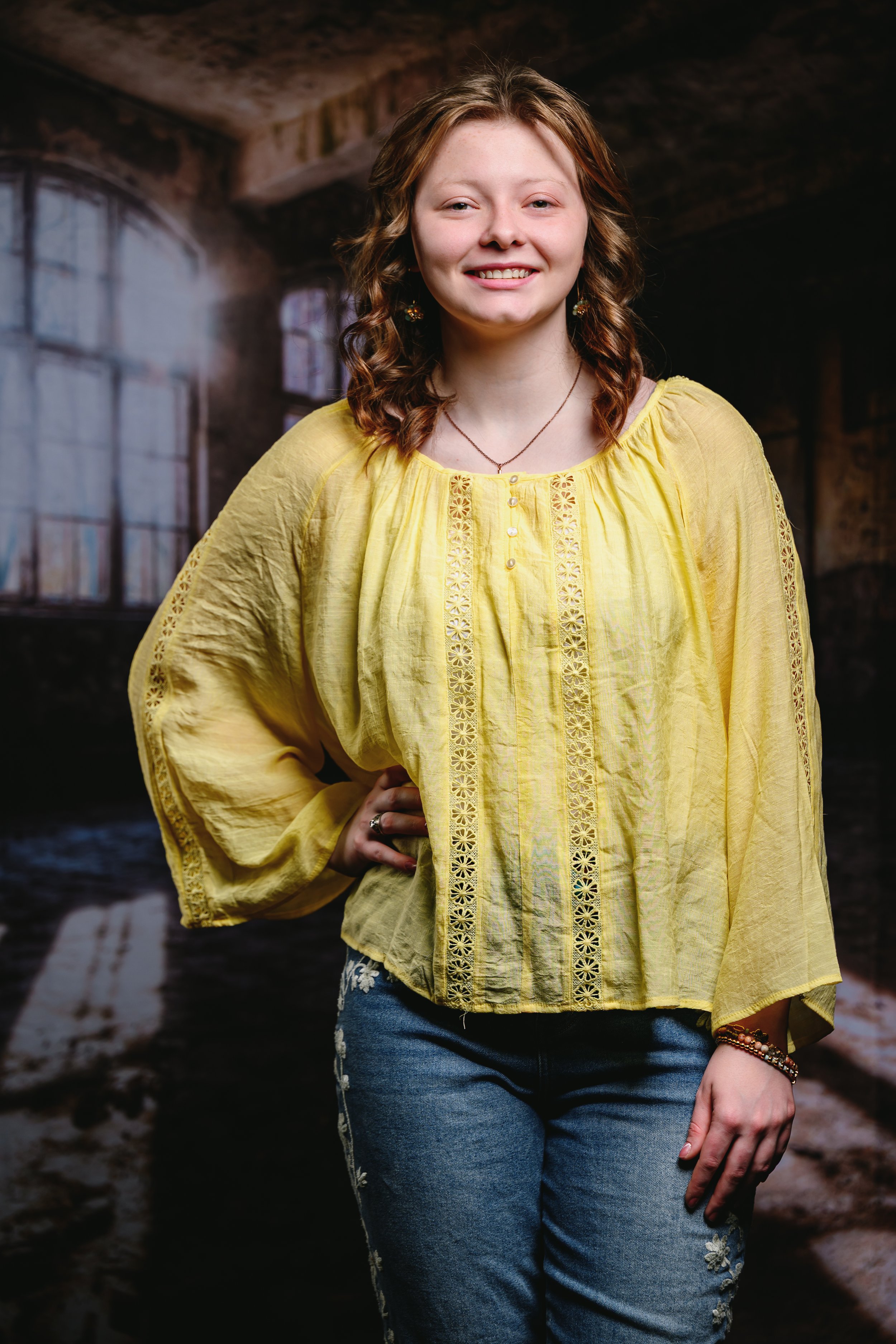 A smiling woman in a yellow blouse and jeans standing indoors with a blurred window background.