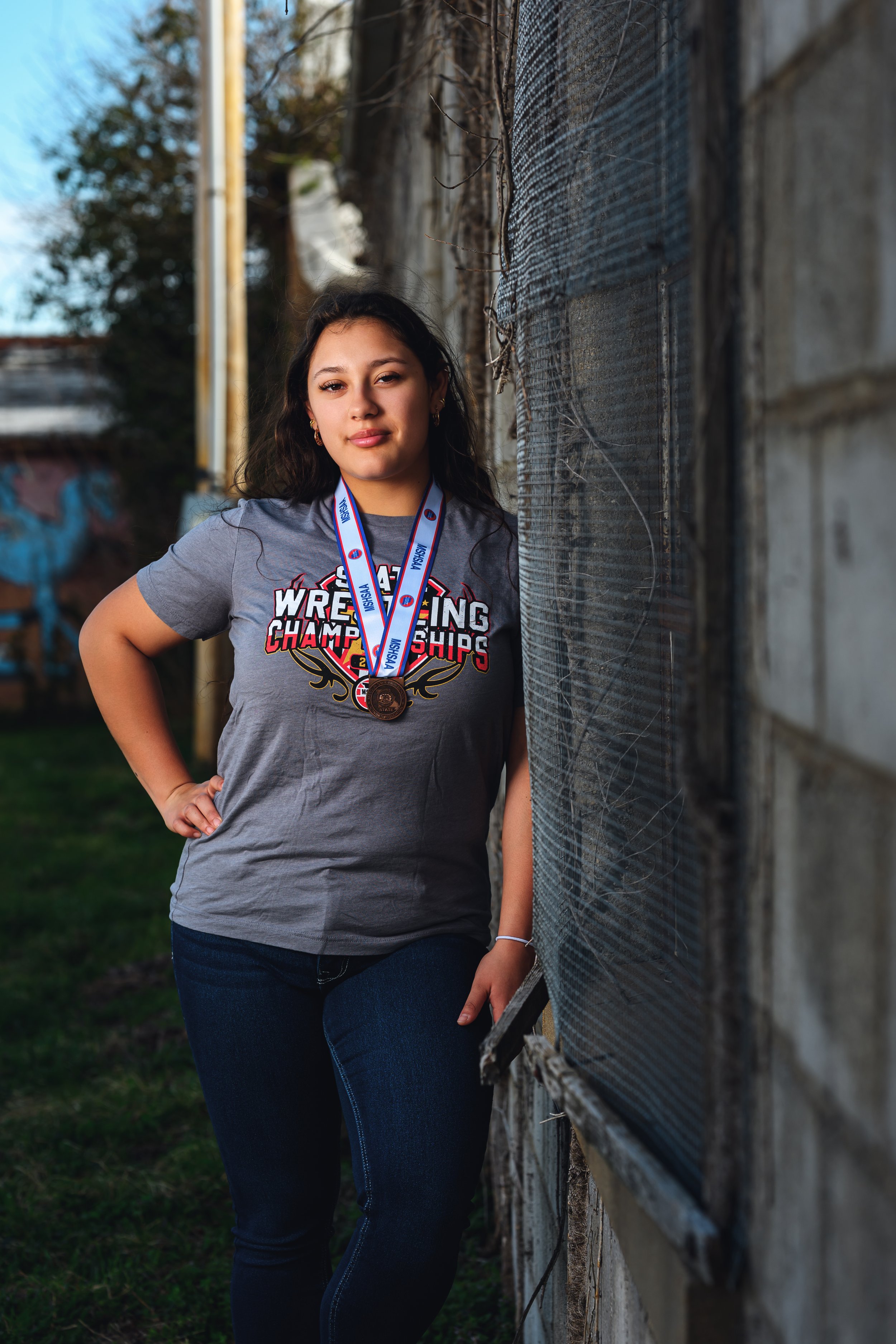 Young woman wearing a wrestling championship t-shirt and medal, standing outdoors next to a wall.